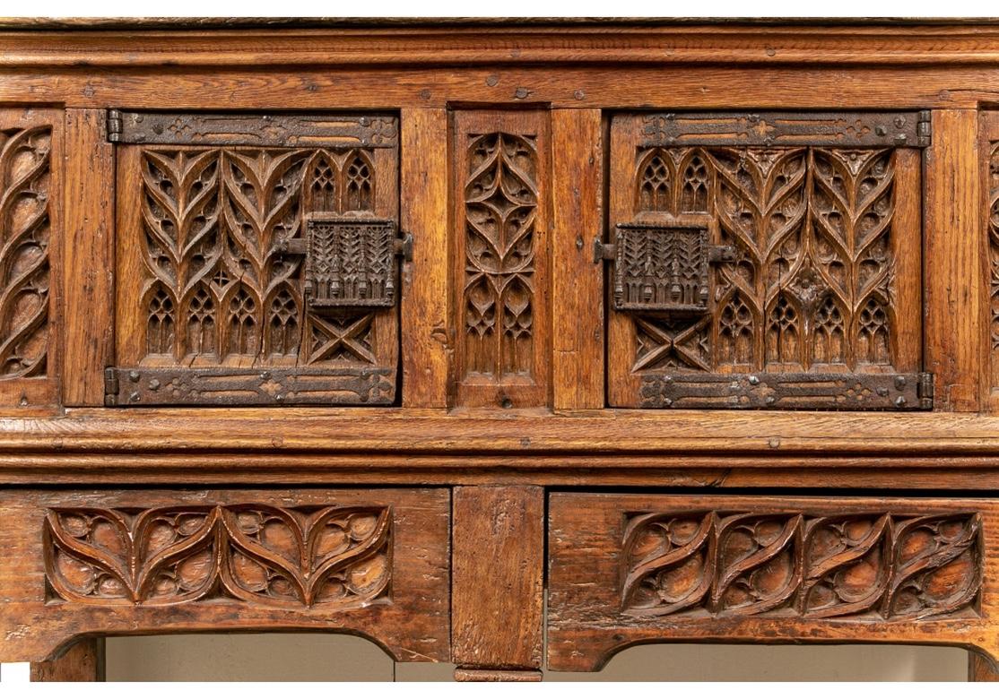An important Neo-Gothic style carved cabinet with carved bands with scrolling vines and pointed windows. The upper double doors with complementary iron hardware over two carved frieze drawers. Recessed and carved side panels. Raised on heavy square