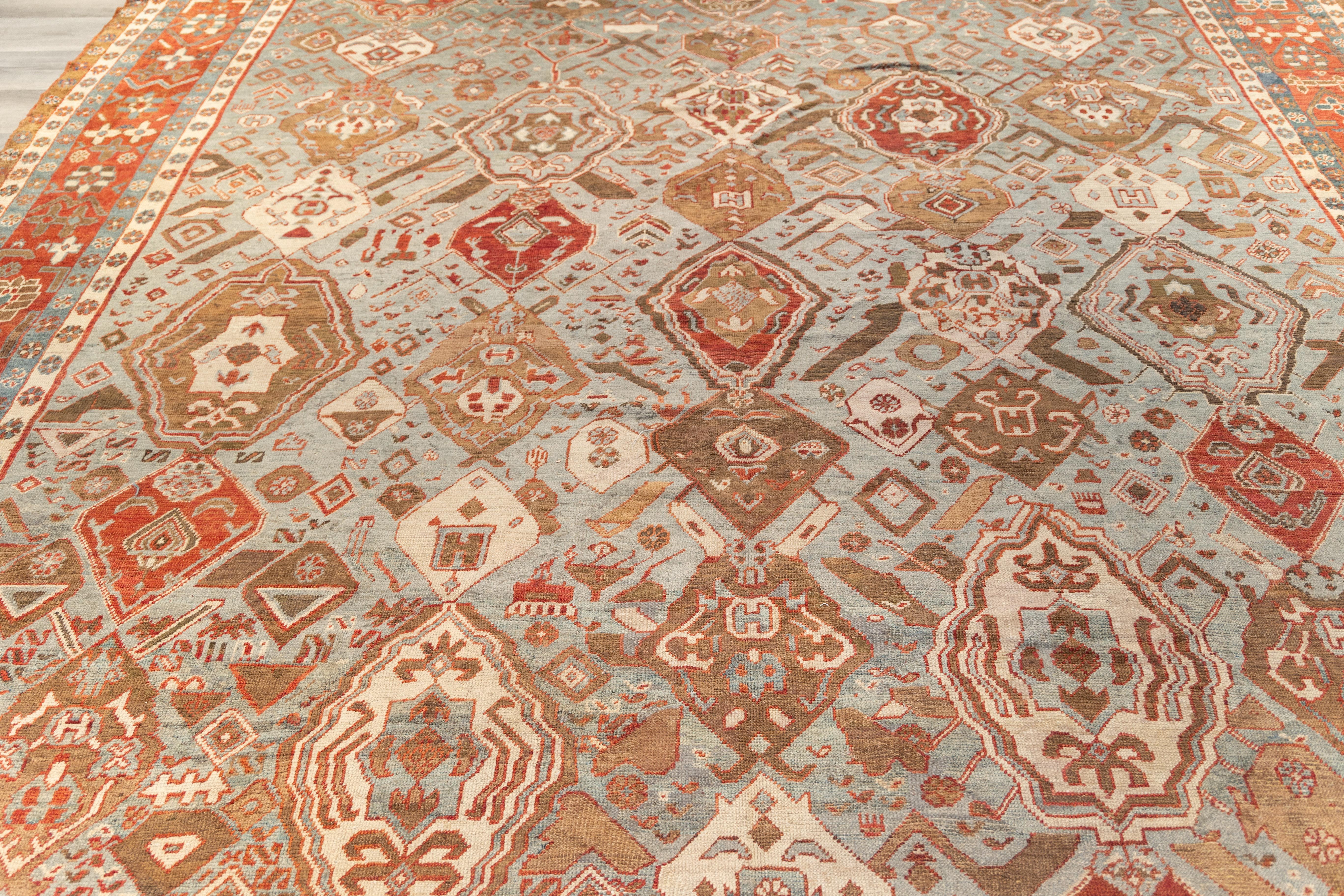 Large Bakhshayesh rug with an expansive field of scattered shields and tribal motifs in a spacious, asymmetric layout. Earthy rust, sand, and ivory tones unfold over a cool, oxidized blue-green background, framed by a wide brick-red border filled