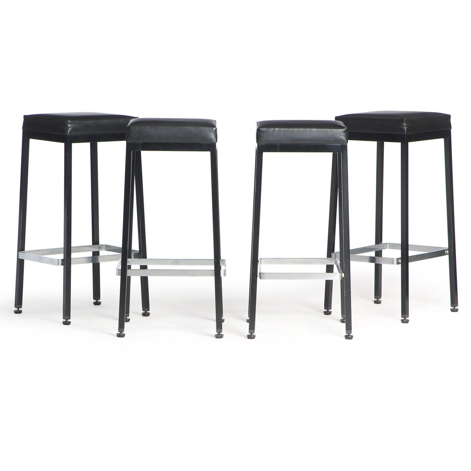 Tabouret de bar ou de comptoir ayant une structure en acier patiné avec un repose-pieds flottant en acier satiné et une assise carrée rembourrée.
