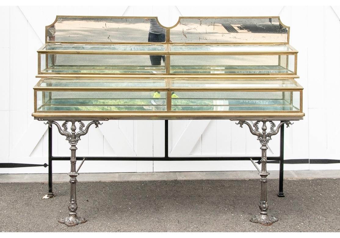 A fine and rare two tiered display stand with fine brass banding, clear glass tops and sides, and mirrored backs. With a shaped top mirrored backsplash over a top tier with long mirror shelf and two hinged sloping clear glass lids. A matching lower