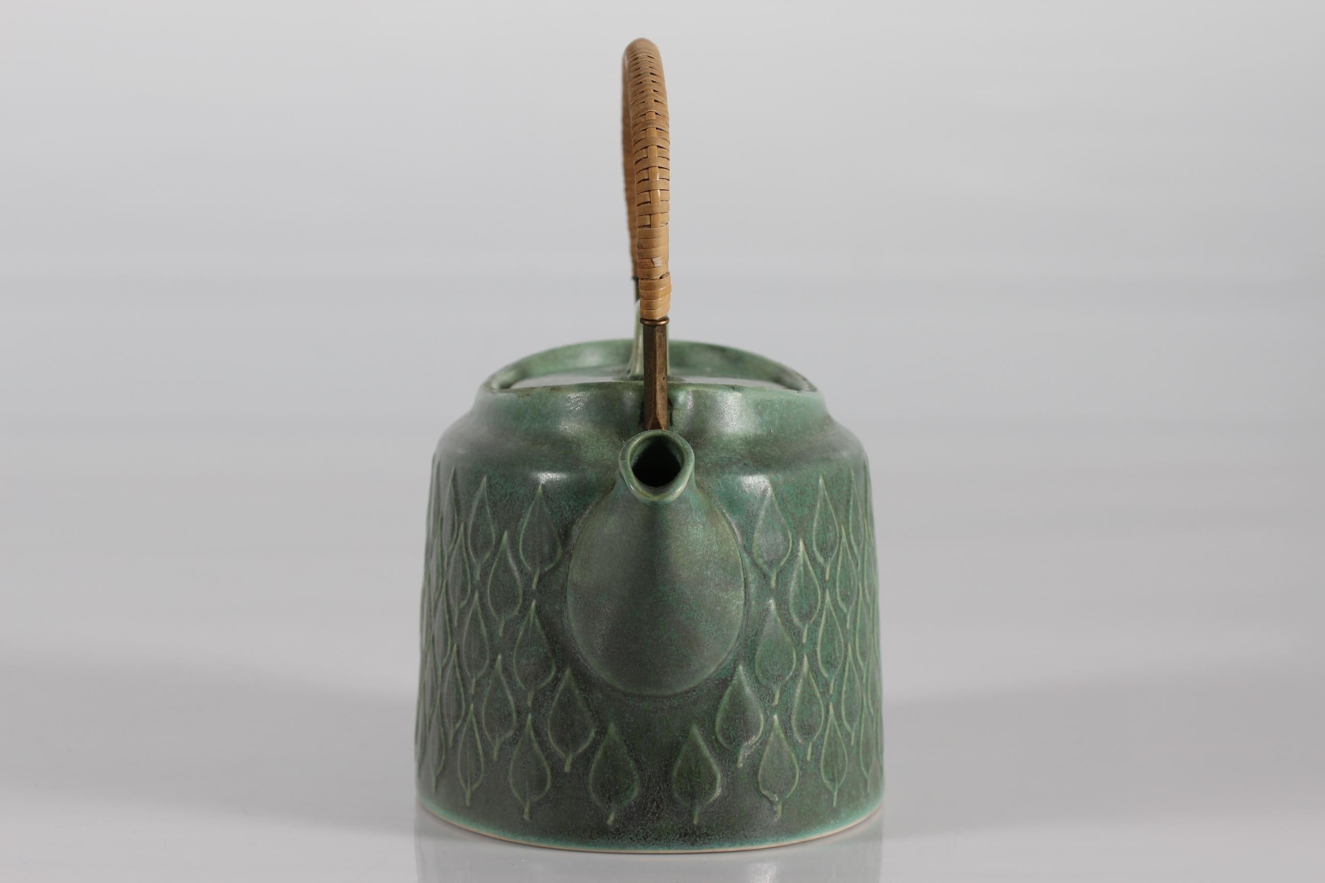 Rare and very early tea pot from the well known service RELIEF launched in 1959 designed by Danish Jens Harald Quistgaard IHQ.

The tea pot has a brass + rattan handle and an embossed leaf shaped decoration. The color of the glaze is salt