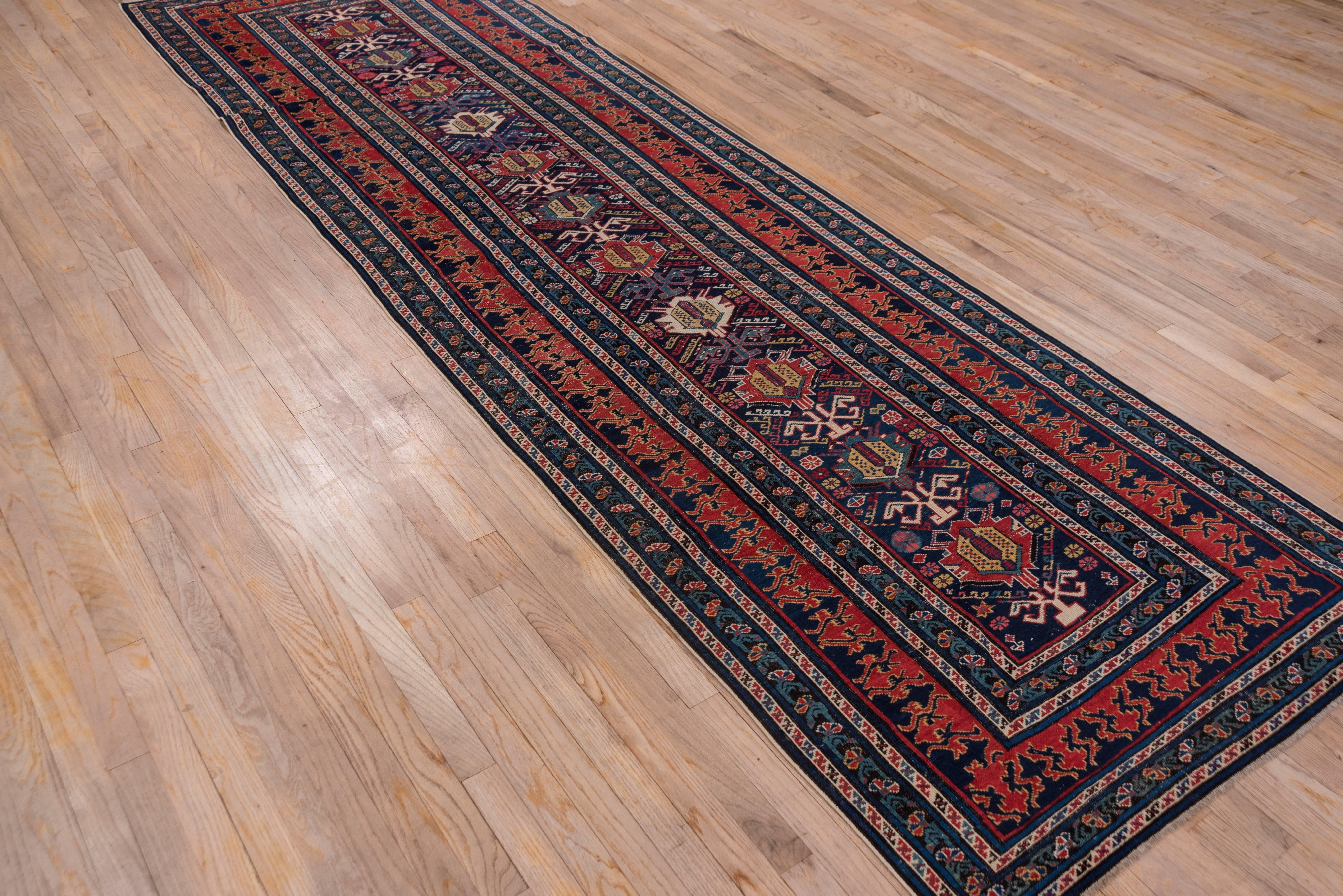 This finely woven east Caucasian long rug displays a field with a one way palmette pattern in red, ivory, green and yellow. The main border is patterned with a complex two colored reciprocal in red and navy. In addition there are two minor stripes