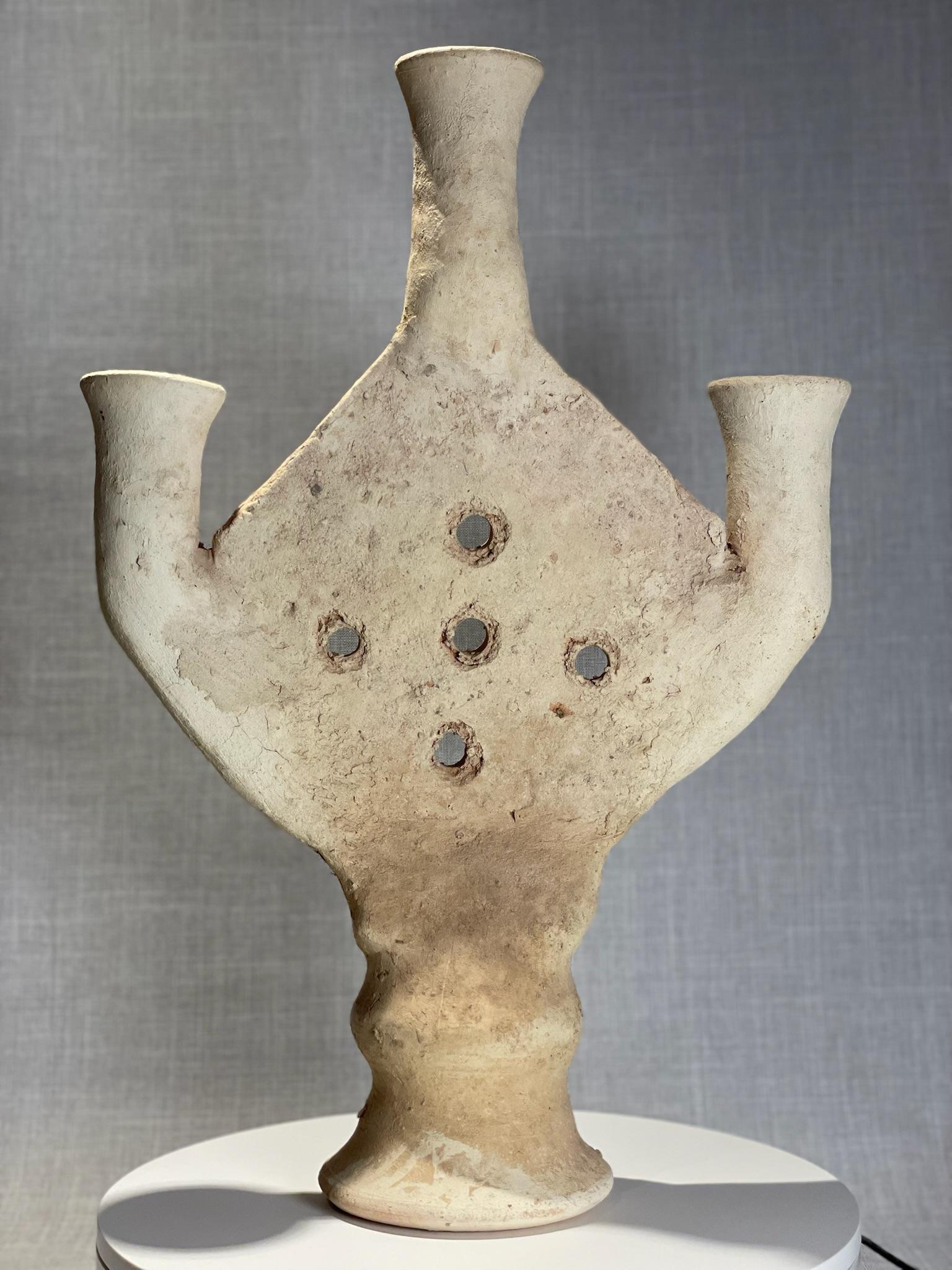 Questo notevole vaso antico in terracotta è uno straordinario esempio di ceramica a più beccucci, realizzata con intenti sia funzionali che simbolici. Poggiato su una robusta base a piedistallo, il vaso si espande in un corpo arrotondato prima di