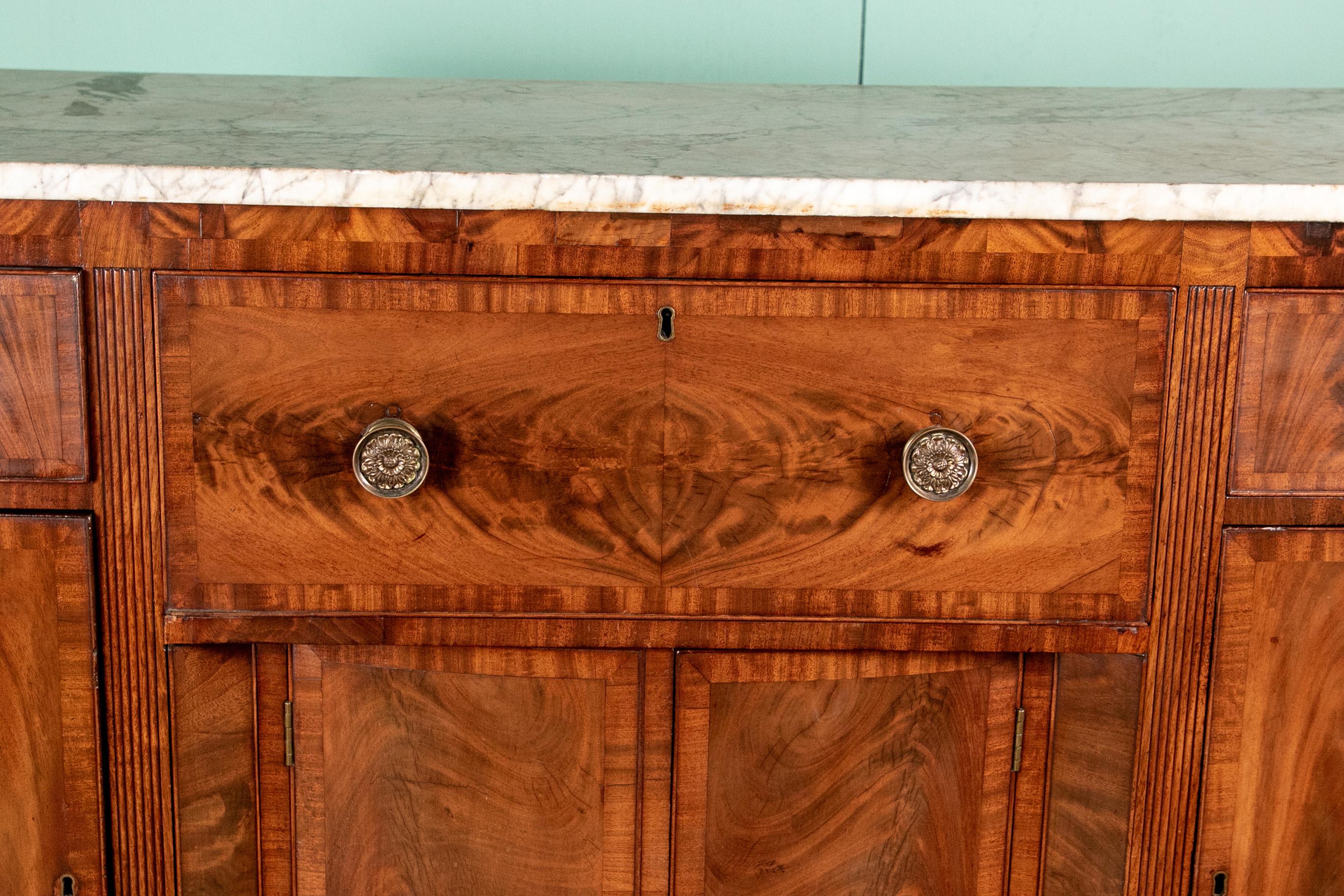 A truly notable antique sideboard/ desk with banded and bookmatched mahogany door and drawer fronts, foliate brass pulls and original gray streaked white marble top. The central top drawer pulls out and folds down to reveal a green felt writing