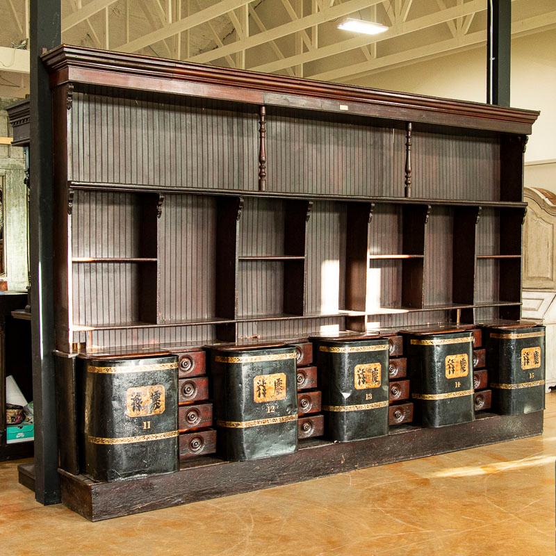 This impressive store display cabinet at over 13' long and 8' high is a rare and unusual find. The wood cabinetry and shelving was made in Scotland and shipped to London to be fitted for the Chinese characters/tole work added to the tin storage