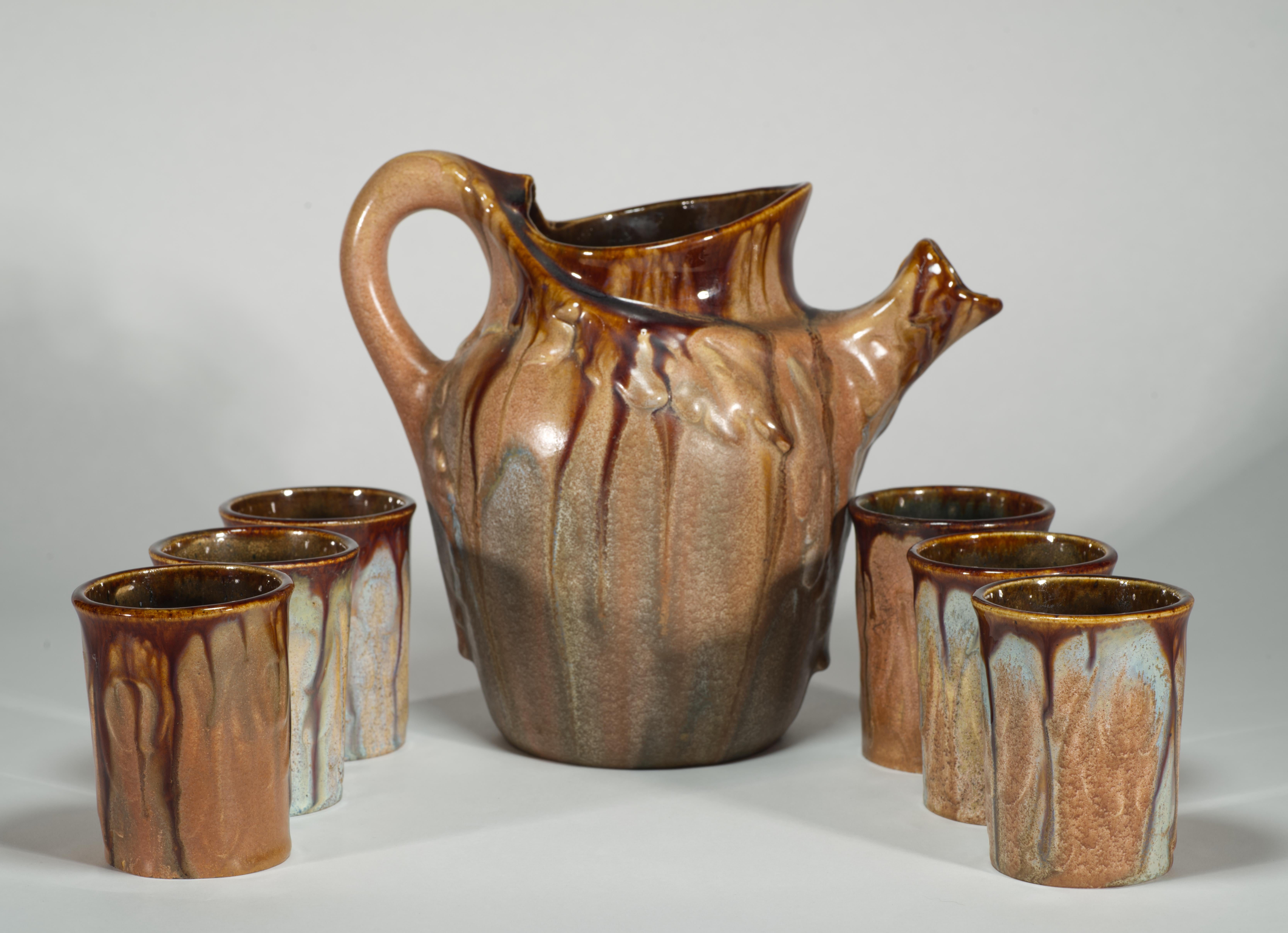  Rare set of a pitcher and 6 glasses or cups was created in characteristic Belgian Art Nouveau style with the use of warm, earthy tones and varied glaze finish on lightly textured shapes of the pieces. Predominantly brown-beige palette is