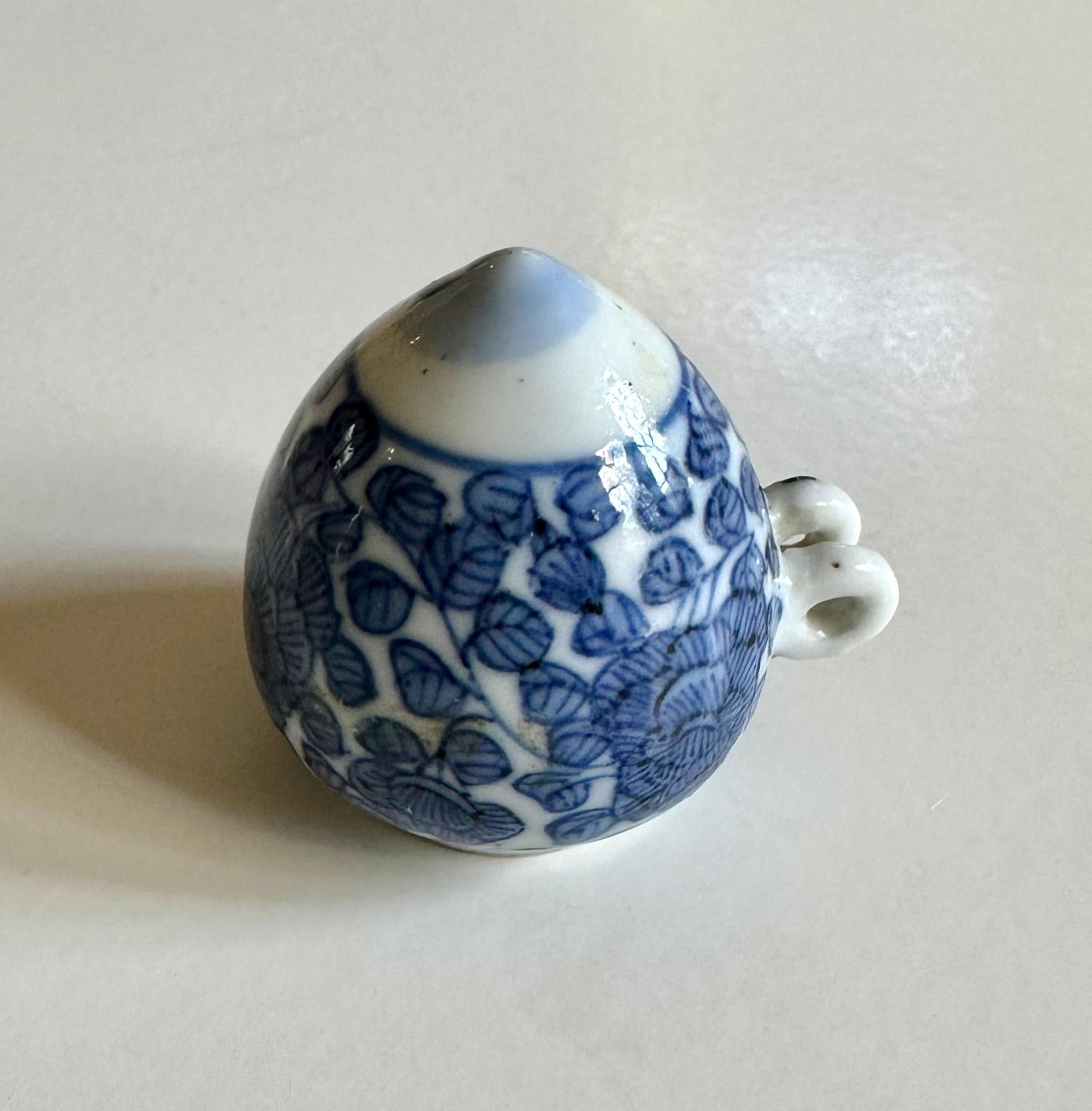 A small blue and white bird-feeder of circular form with short neck, one side set with two small loup handles, the base slightly pointed, painted with lotus heads growing from scrolling stems with curled leaves between a double-line border