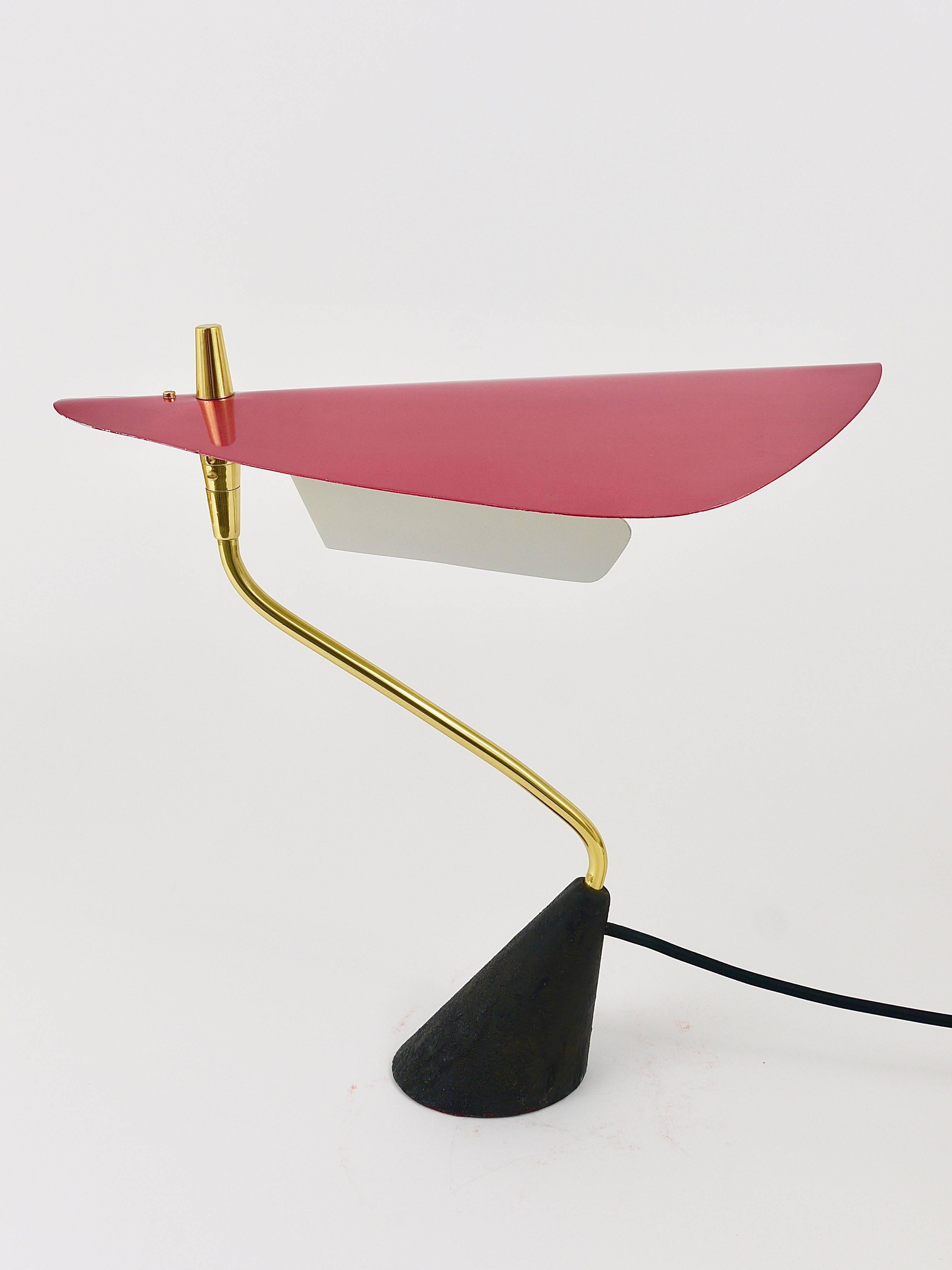 A sculptural table or desk lamp from the 1950s, executed by Werkstätte Karl Hagenauer. Has a conical black patinated cast-iron base, an asymmetrical brass tube and a red anodized aluminium lampshade with white lacquered interior. The lampshade is