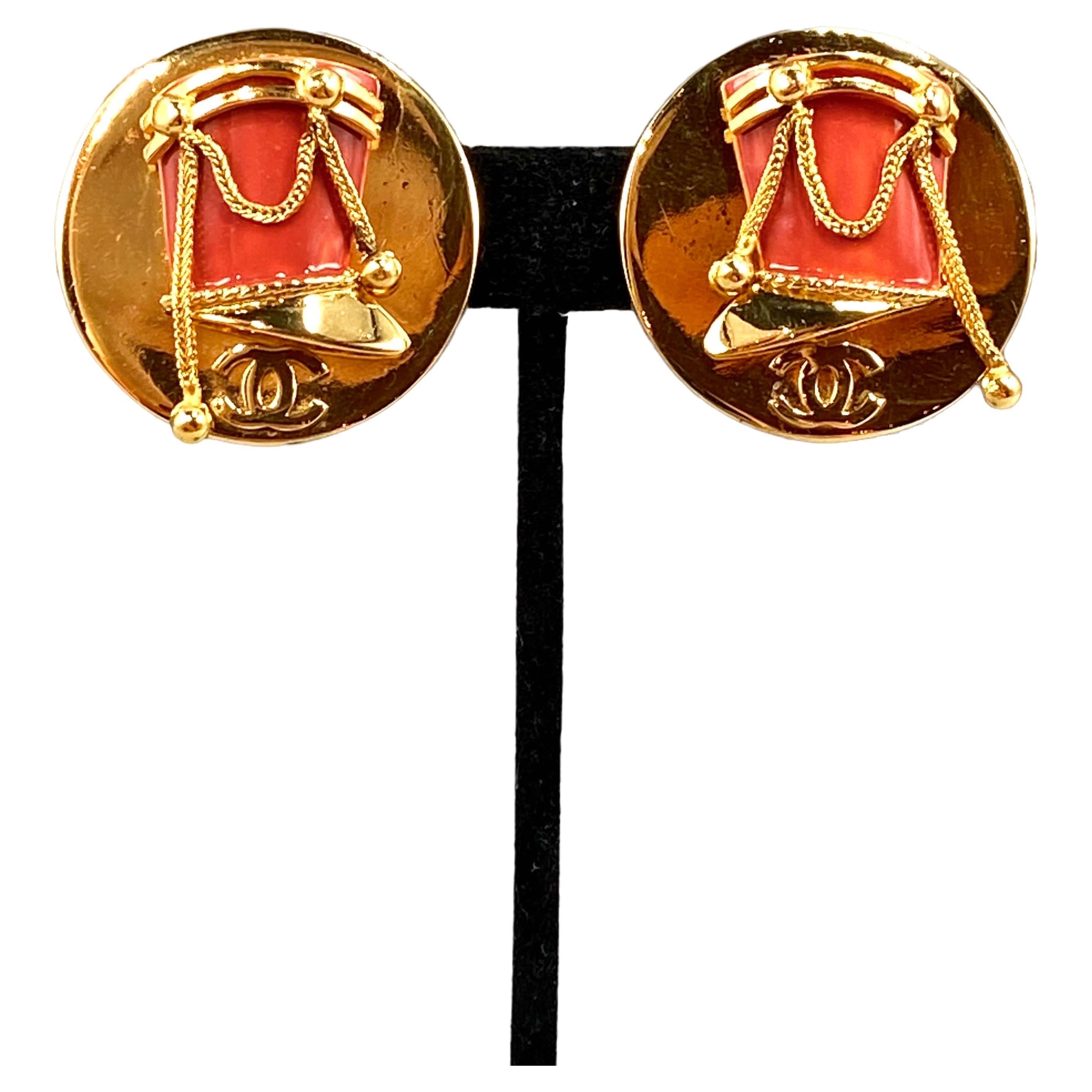 A rare pair of large Chanel button earrings from the early 1970s. Each measures 1.5 inches in diameter and features a three dimensional French military hat. The hat is accented with red enamel and free hanging gold chain swag with ball ends. Below