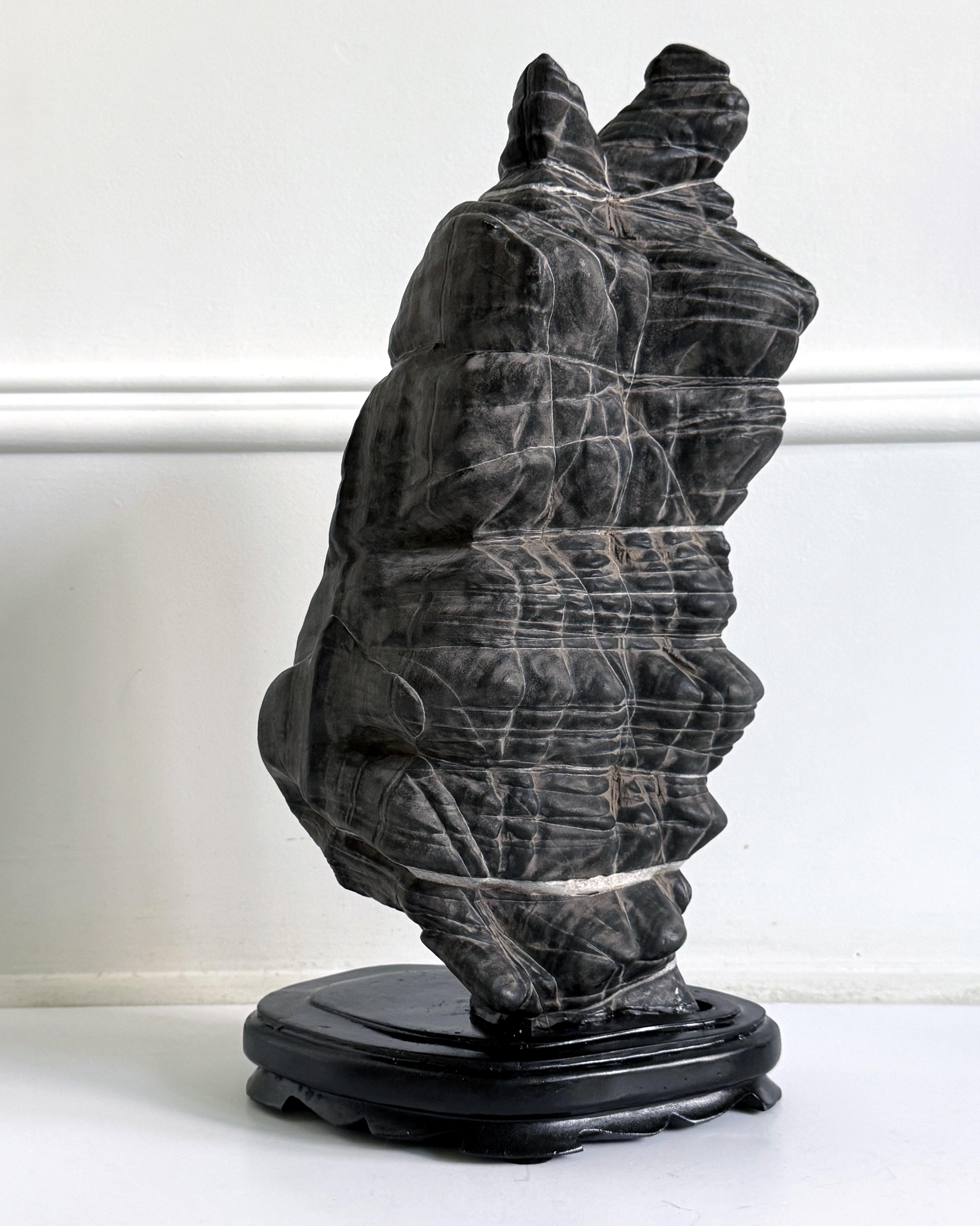 A Chinese scholar stone of Lingbi type presented on a carved wood base. This stone takes a singular vertical form with one convex side that is largely flat, and the other side slightly concaved with prominent folding. It is supported by a small