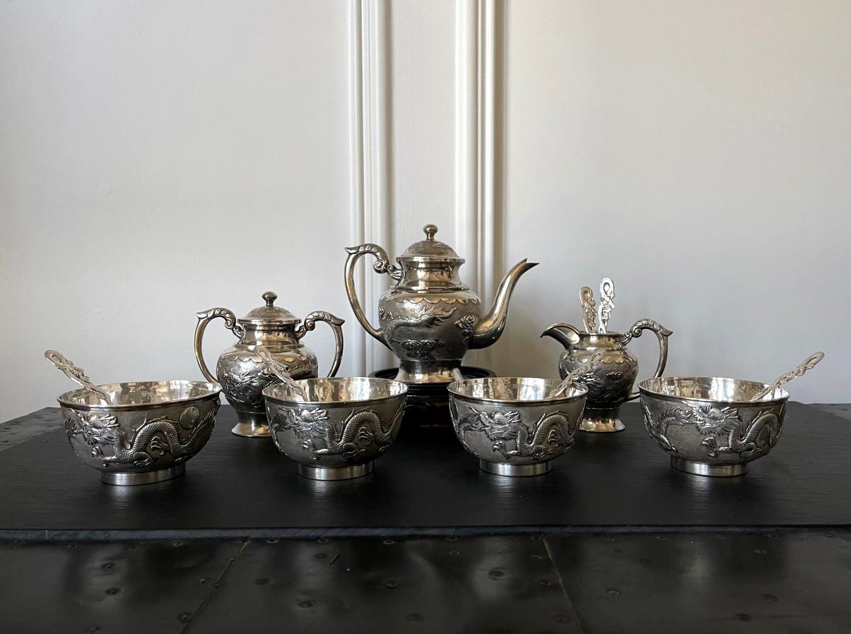 A Chinese export sterling silver tea set comprised of 13 pieces including tea pot, sugar, creamer, four tea bowls and six tea spoons, circa 1910s-1920s. Featuring matching chased relief dragon and cloud motif on a contrasting tightly hammered