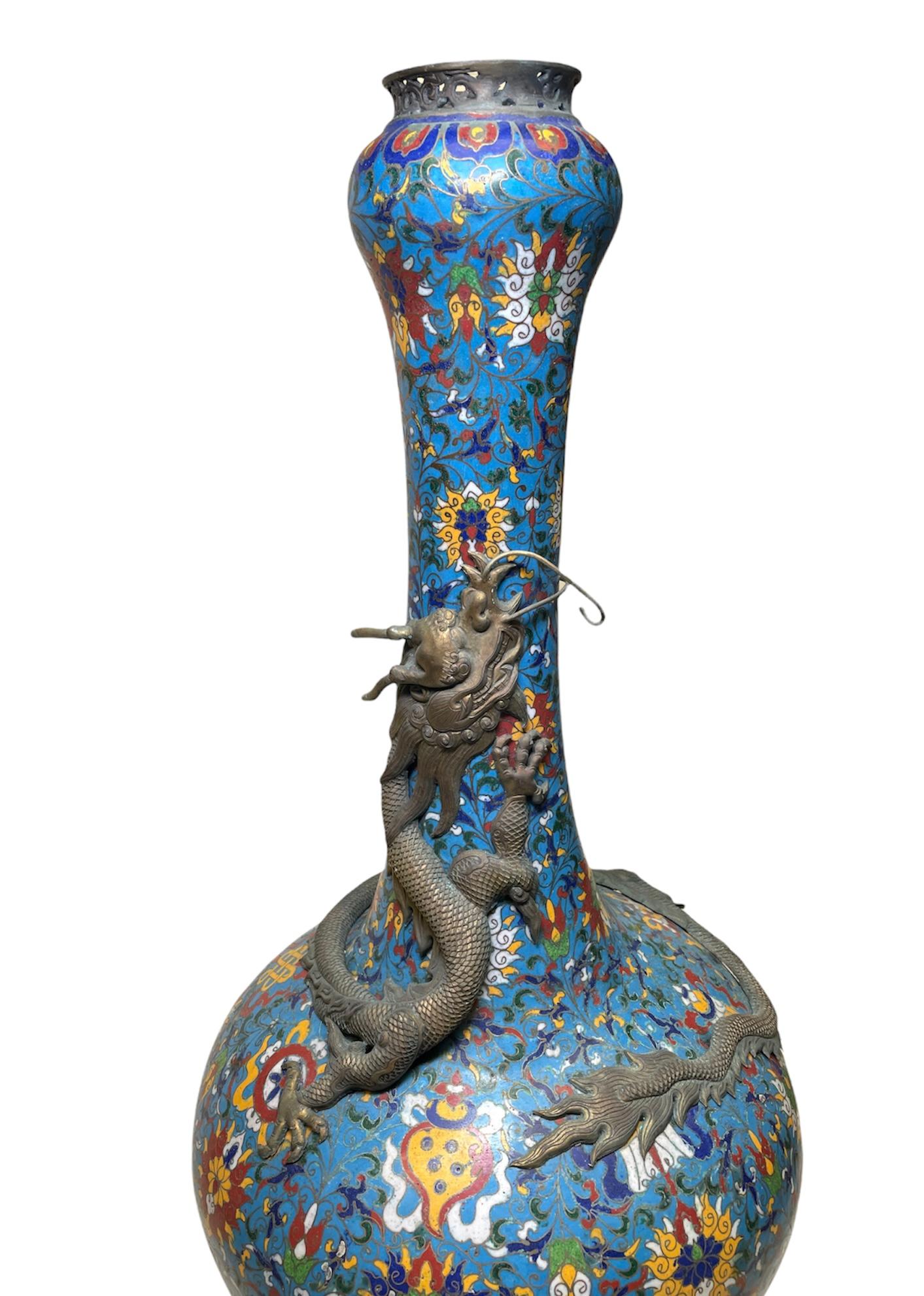 This is a Chinese colorful cloisonné large vase with long neck and round belly. It has a turquoise background adorned with different flowers, scrolls leaves and Chinese knots in multiple bright colors. There is a large bronze dragon that has already