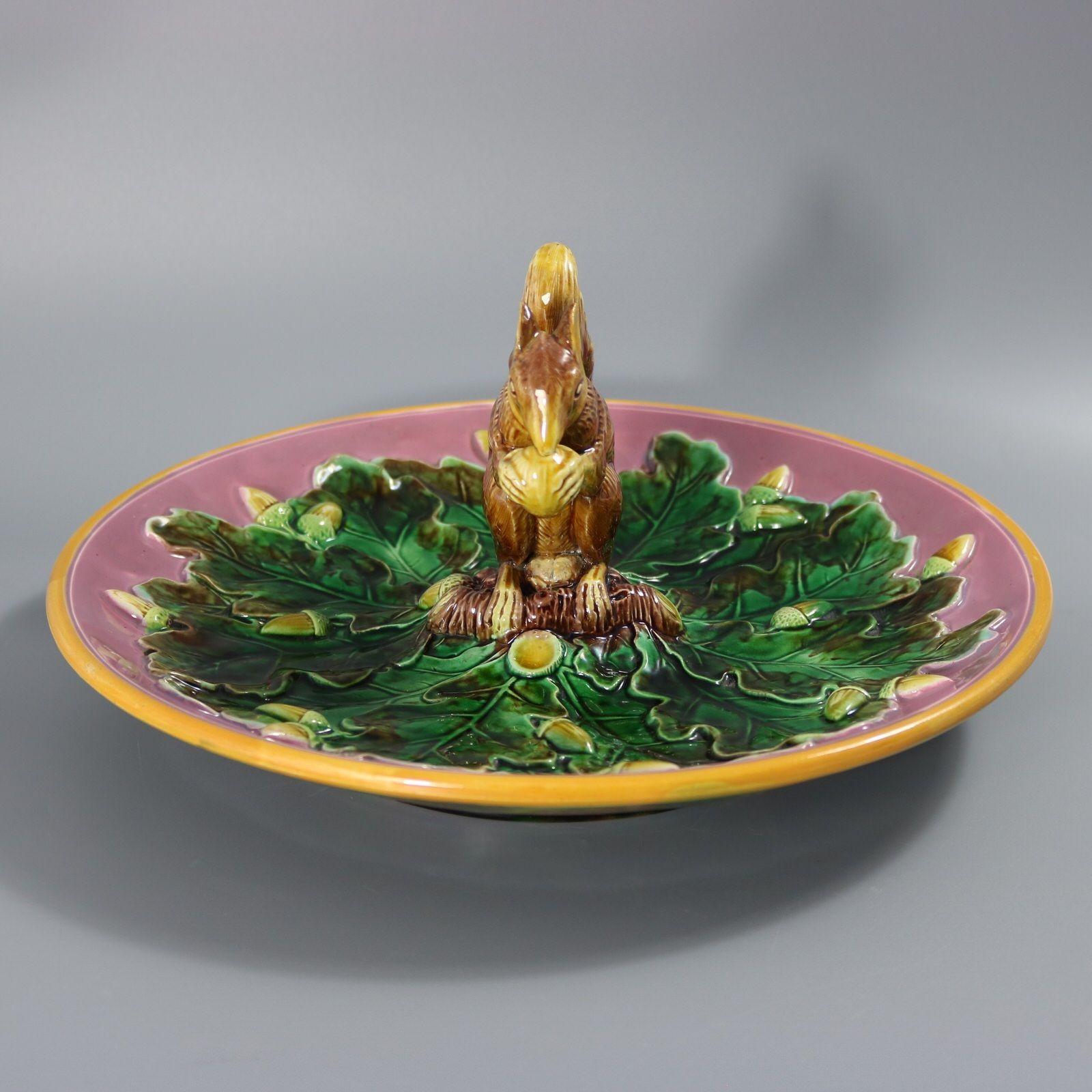 Piatto in maiolica di George Jones con uno scoiattolo seduto su un ramo che tiene in mano una noce. Circondato da ghiande e foglie di Oak. Versione a terra rosa. Colorazione: predominano il verde, il rosa e il marrone. Porta un numero di modello,
