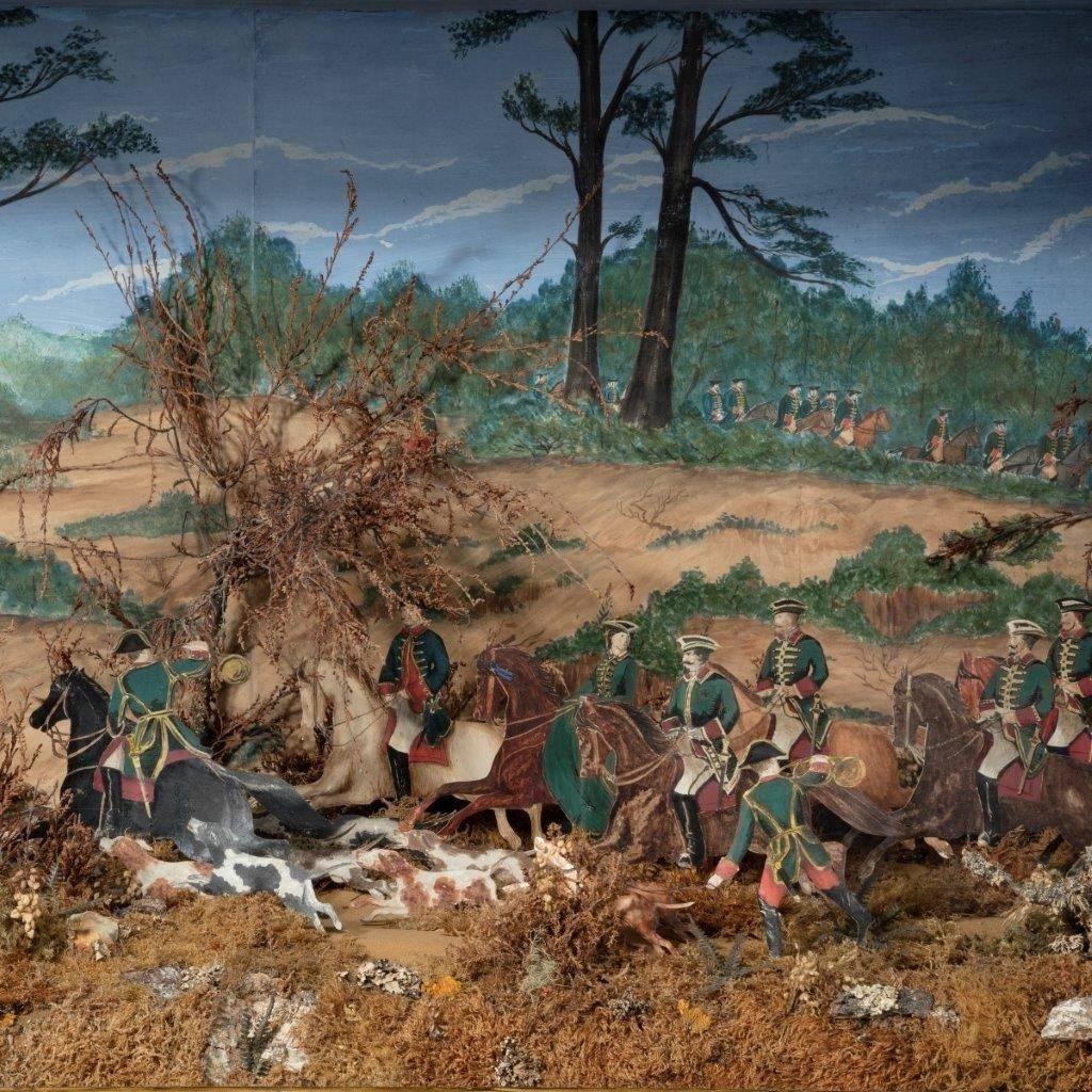 A rare Continental three dimensional cut paper diorama showing a band of green liveried huntsmen chasing a stag in a wooded landscape, in a mahogany show case, circa 1850. On a later stand.