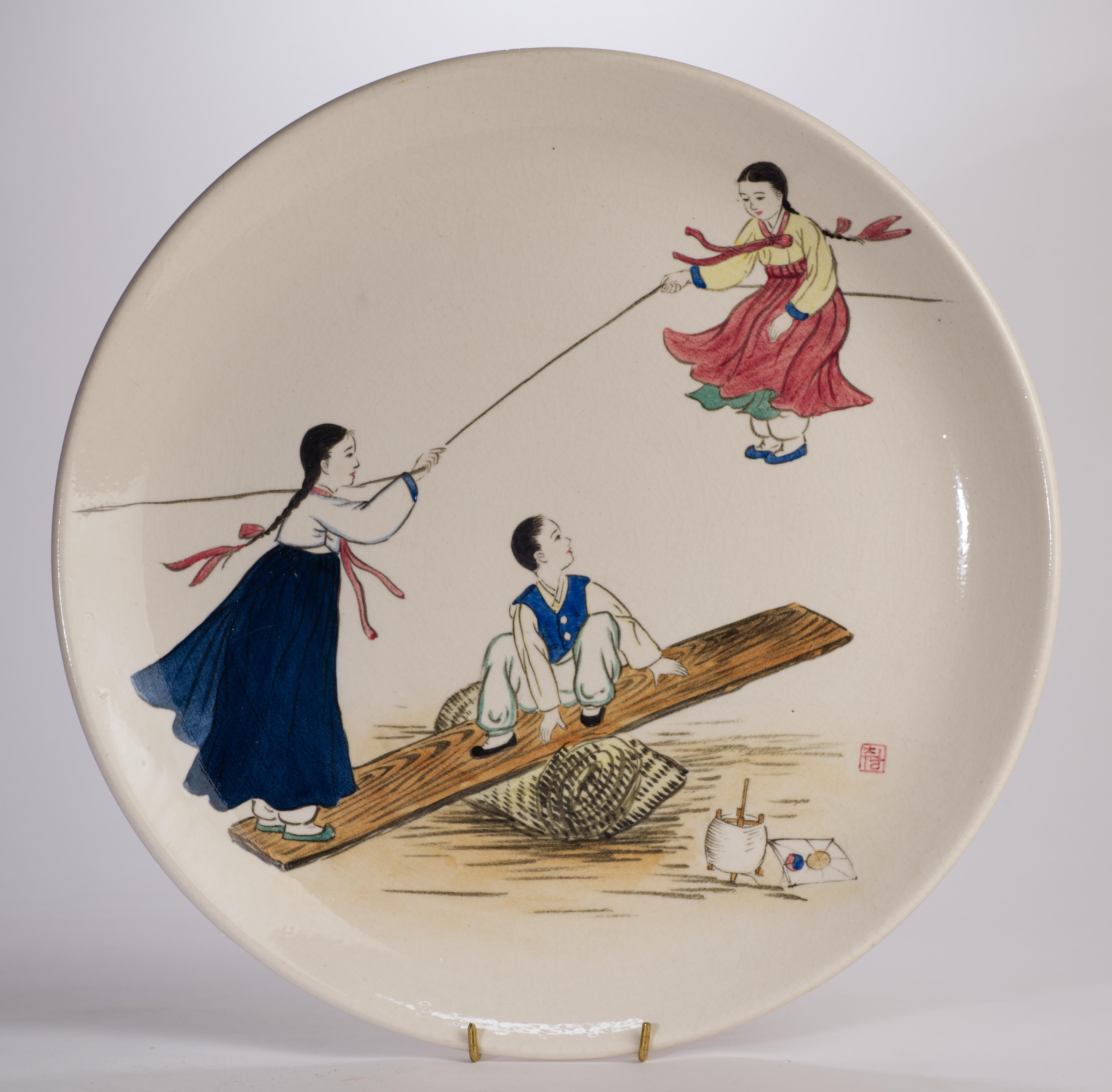  El plato de cerámica decorativa vintage está decorado con una escena pintada a mano de dos niñas y un hombre vestidos con trajes tradicionales jugando en un balancín mientras sujetan una cometa, con una pequeña cometa en el suelo junto a ellos.