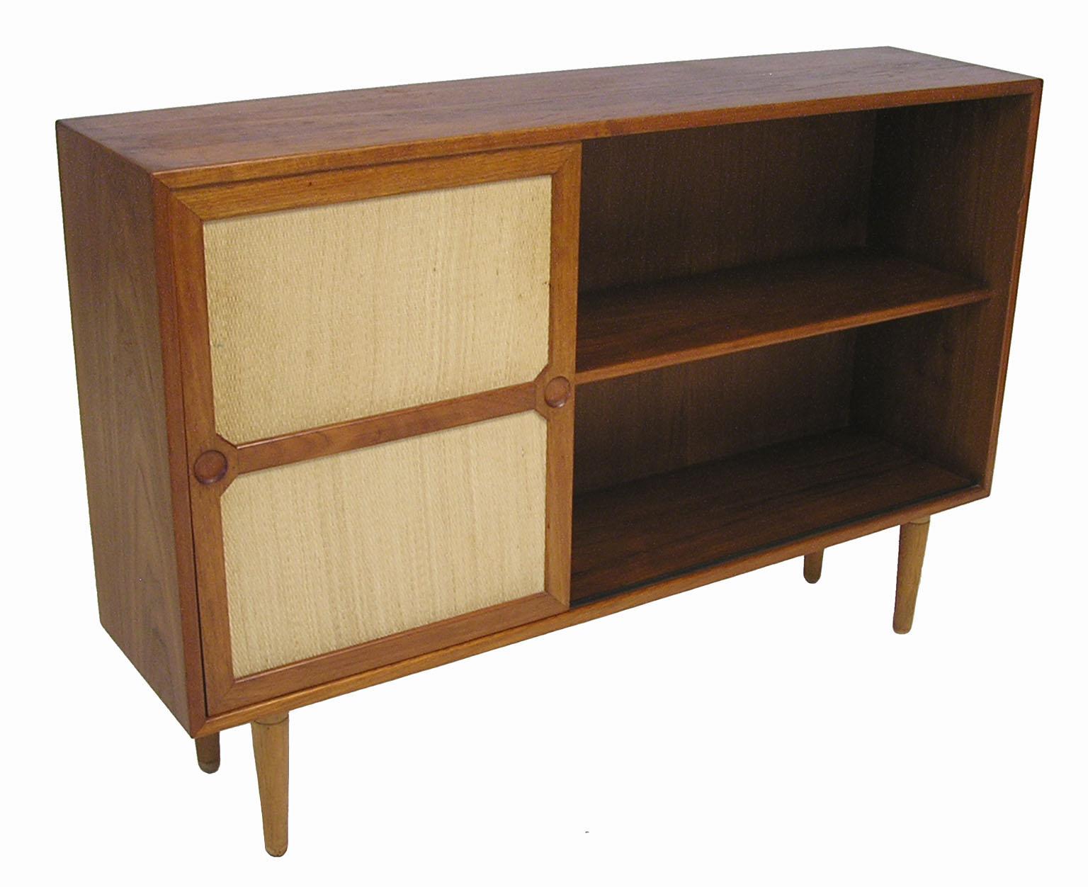 A rare teak bookshelf cabinet from the 1950s Danish Mid-Century Modern era. Quality craftsmanship throughout featuring a unique seagrass panelled sliding door with inset elliptical pulls, adjustable shelving and tapered oak conical legs. The low and