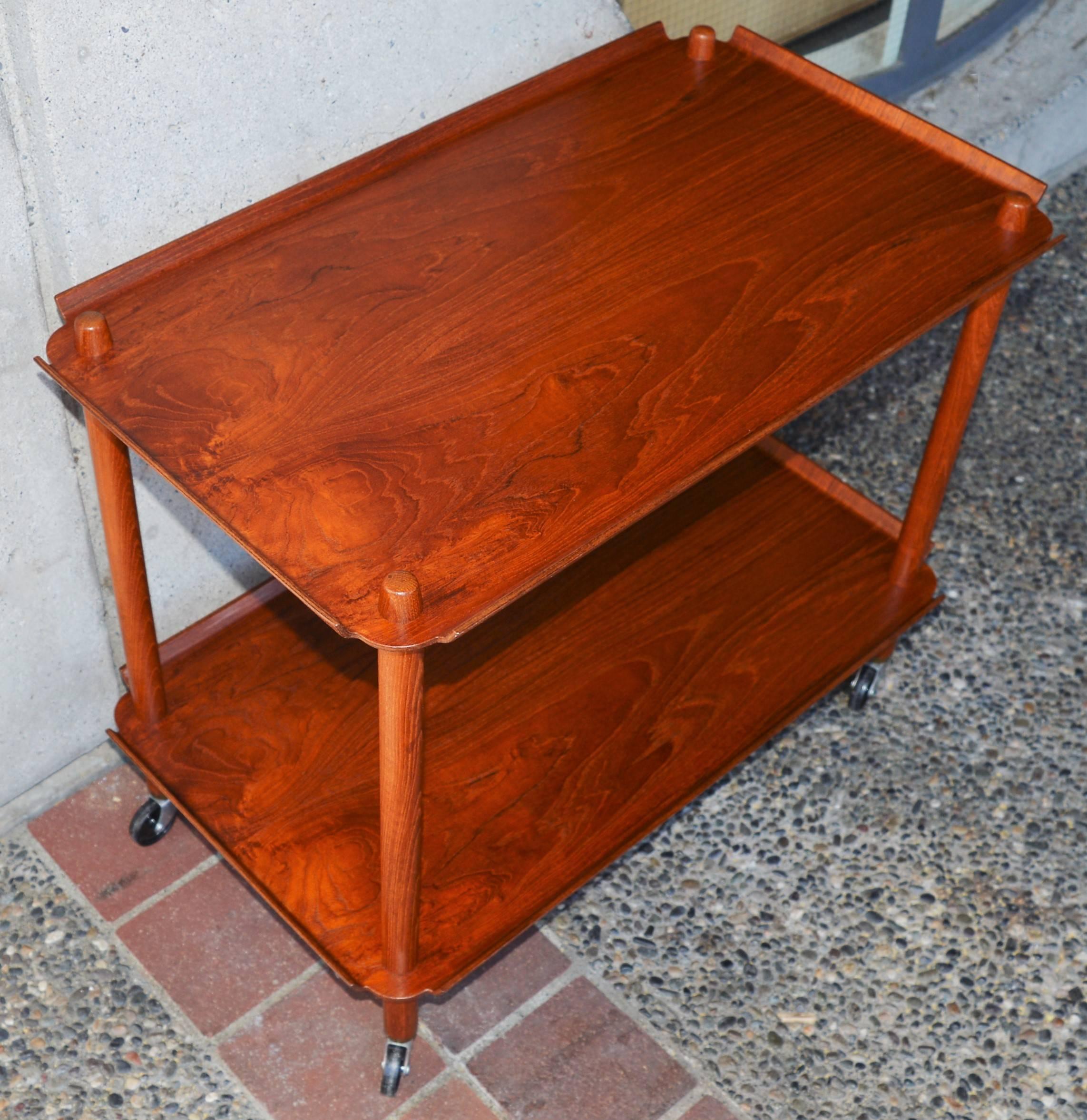 This lovely Danish modern teak two-tier bar cart is a rare version made by Poul Hundevad. Cleverly designed, it can be completely disassembled where it looks like the legs pierce the tiers. Each bentply tier has gorgeous flared edges and the top of