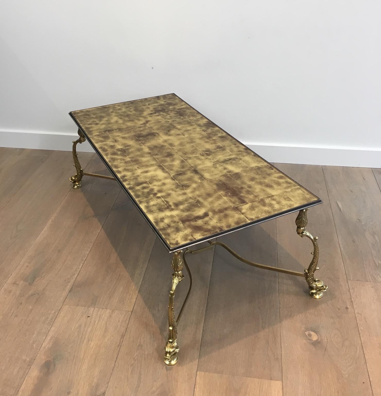 This rare and beautiful neoclassical stytle coffee table is made of brass. It has elegant dolphin feet joined by a stretcher with brass rope on which centre is a decoration. The top is made of a beautiful under glass gold paper marquetry surrounded