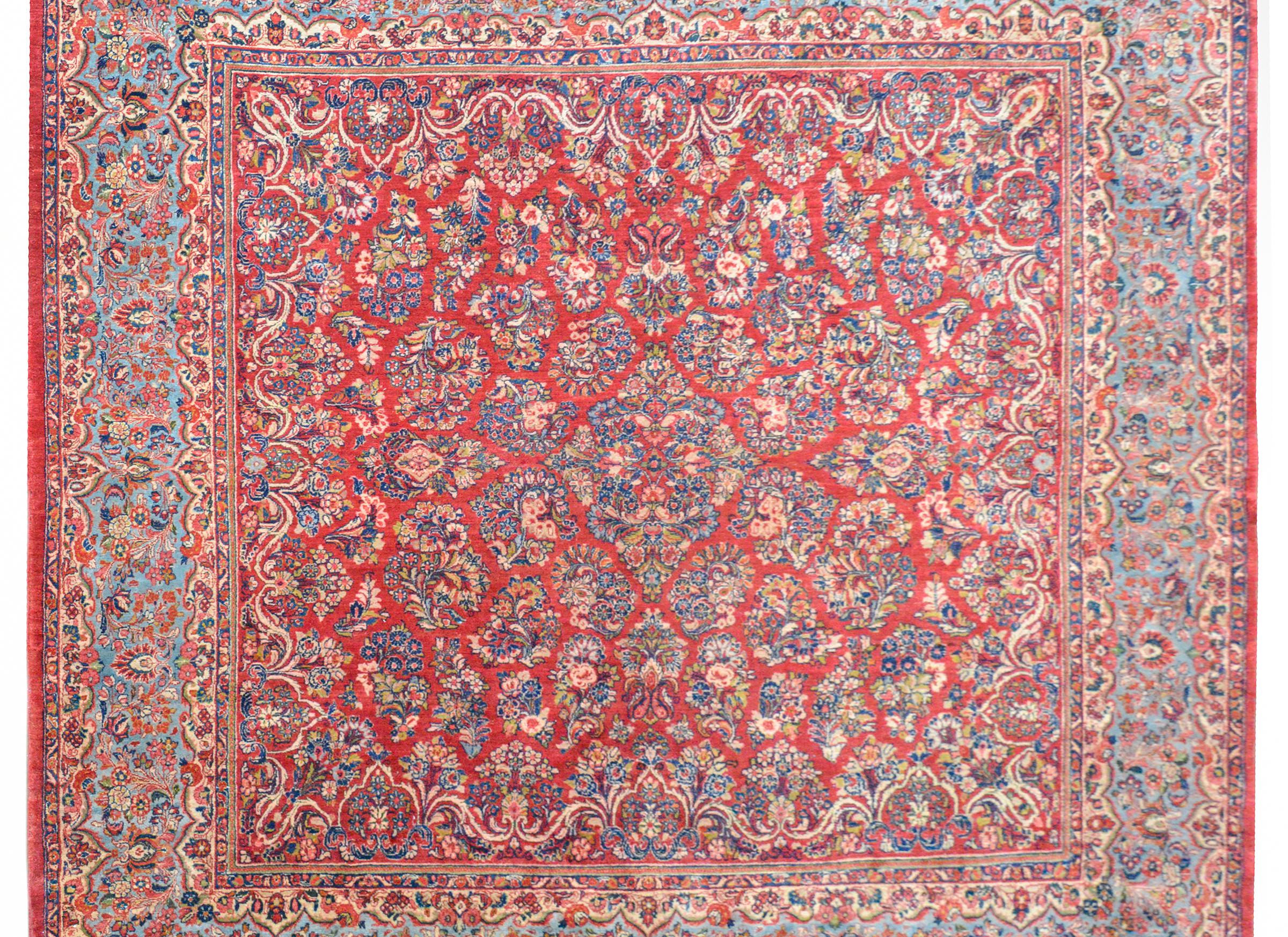 A rare early 20th century square Persian Sarouk rug with a traditional pattern containing myriad clusters of flowers woven in light and dark indigo, cream, pink, and green, set against a crimson background. The border is extra wide, with a central