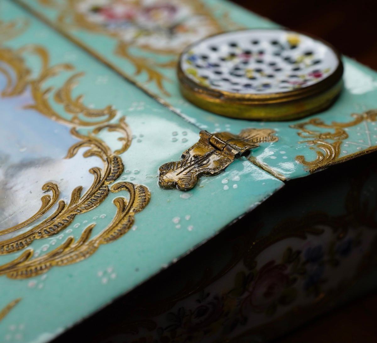English enamel writing box, modelled after a writing slope, with hinged flap and inset sander and inkwell, the top decorated with a large landscape panel within rococo scroll frame, with similar reserves of flowers to the other faces,
circa 1780.