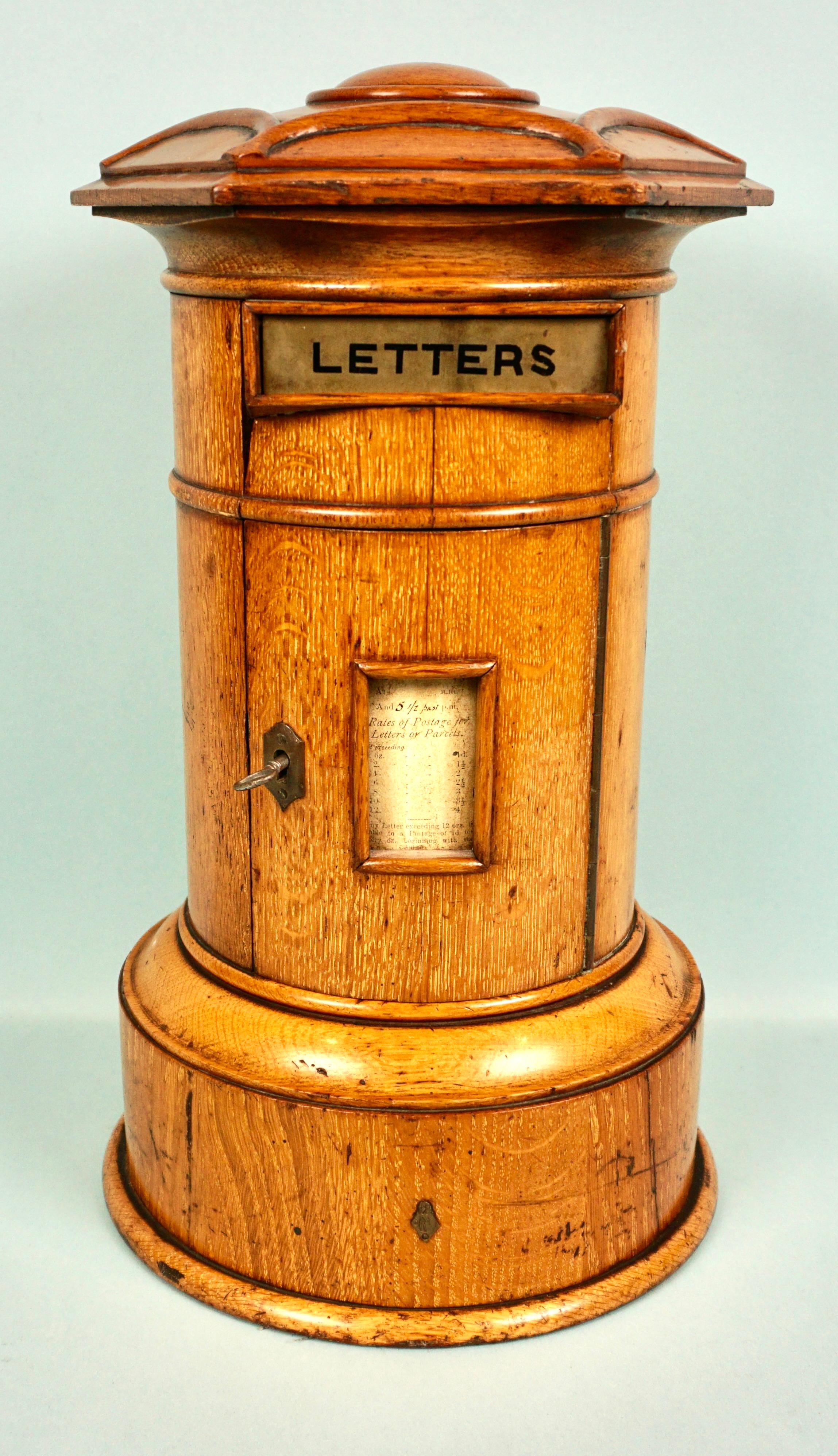 A rare English oak pillar form country house letter box with a circular base and cylindrical body ending in a carved hexagonal lid with a domed top. The front is fitted with a brass opening marked letters, below is a handwritten schedule of pickup