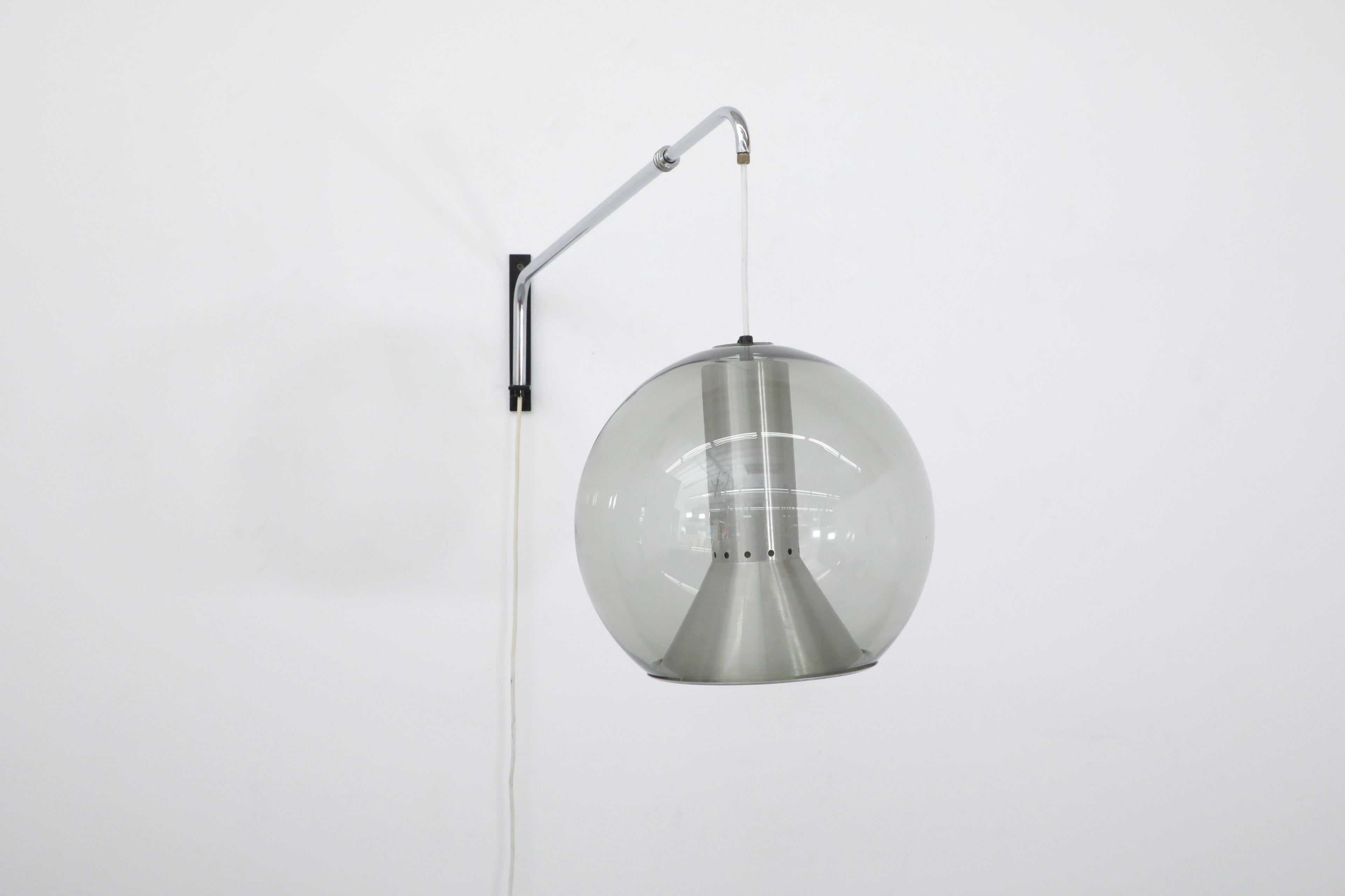Incredible Frank Ligtelijn wall lamp with blown smoked glass globe for Raak. A beautiful rare wall light with extendable and swinging brushed chrome stem on thin black enameled metal mount. There is a trumpet shaped spun aluminum inner shade. The