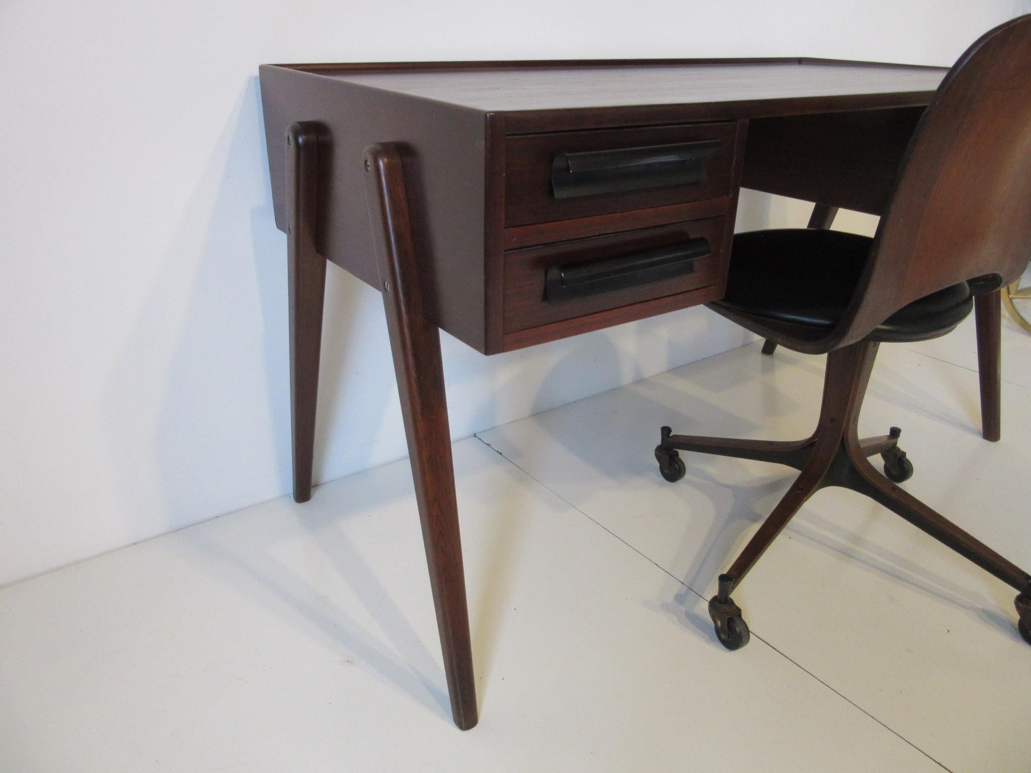 A hard to find Plycraft compass leg desk in a dark walnut tone teak and oak with three drawers, curved black metal pulls and matching sculptural bent ply chair with upholstered bottom pad and wheels . The detail to the desk with it's tapered top
