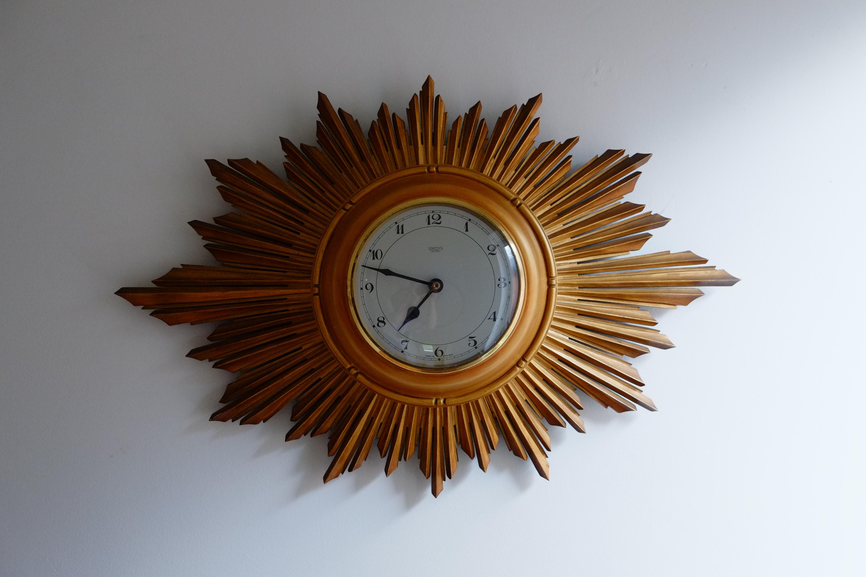 Beautiful 1960s or 70s mid-century wall clock by Smiths clock makers. This rare clock has a stunning large golden oval shaped sunburst, hand carved from wood and a simple aluminium clock face. Made in England. This piece has a particularly elegant