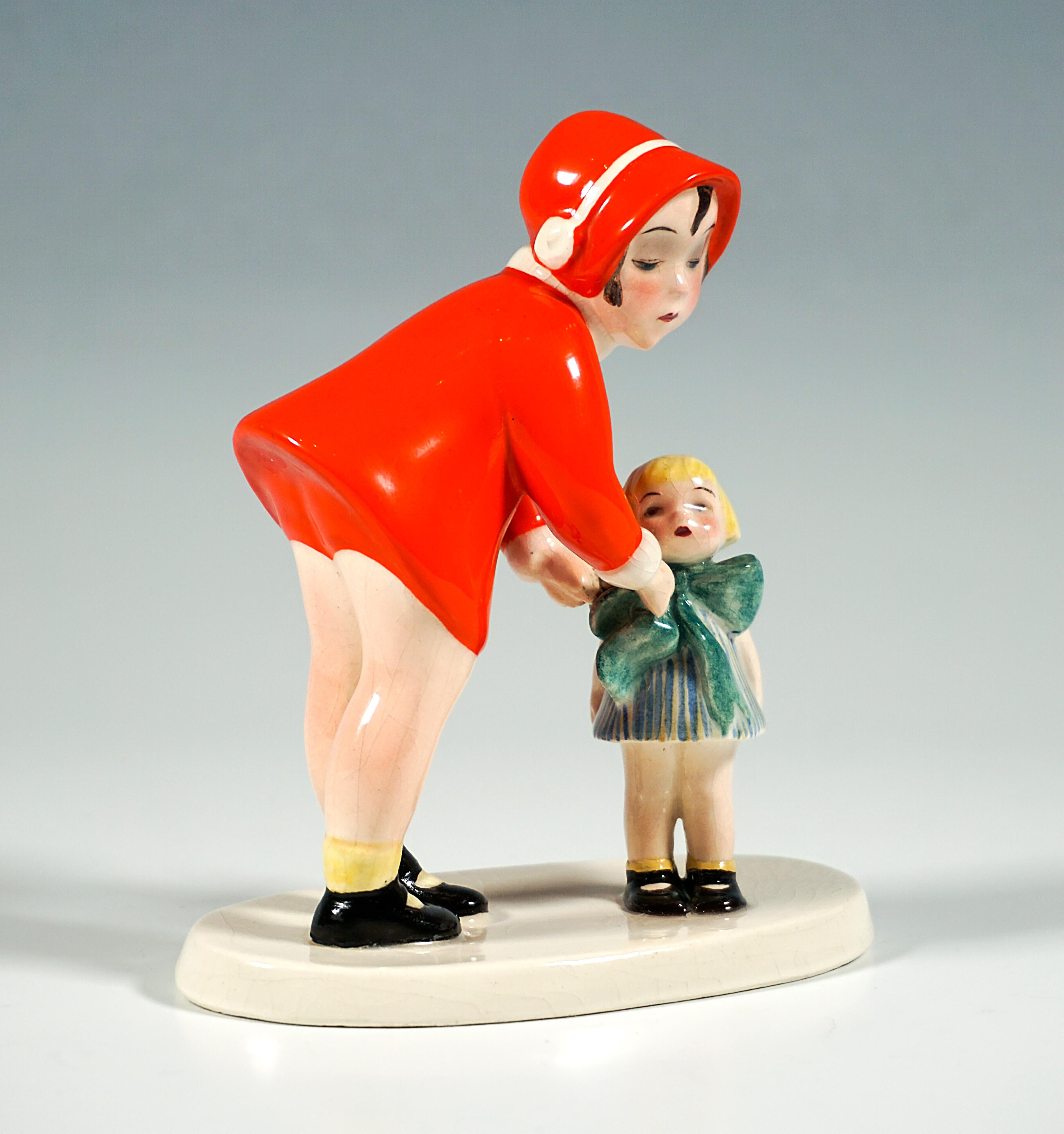 Little girl in a red short coat with a white collar, matching short pants and a red barge hat with a white ribbon, bending down towards the upright doll with a blonde pageboy head and carefully adjusting the large green bow on the collar of the