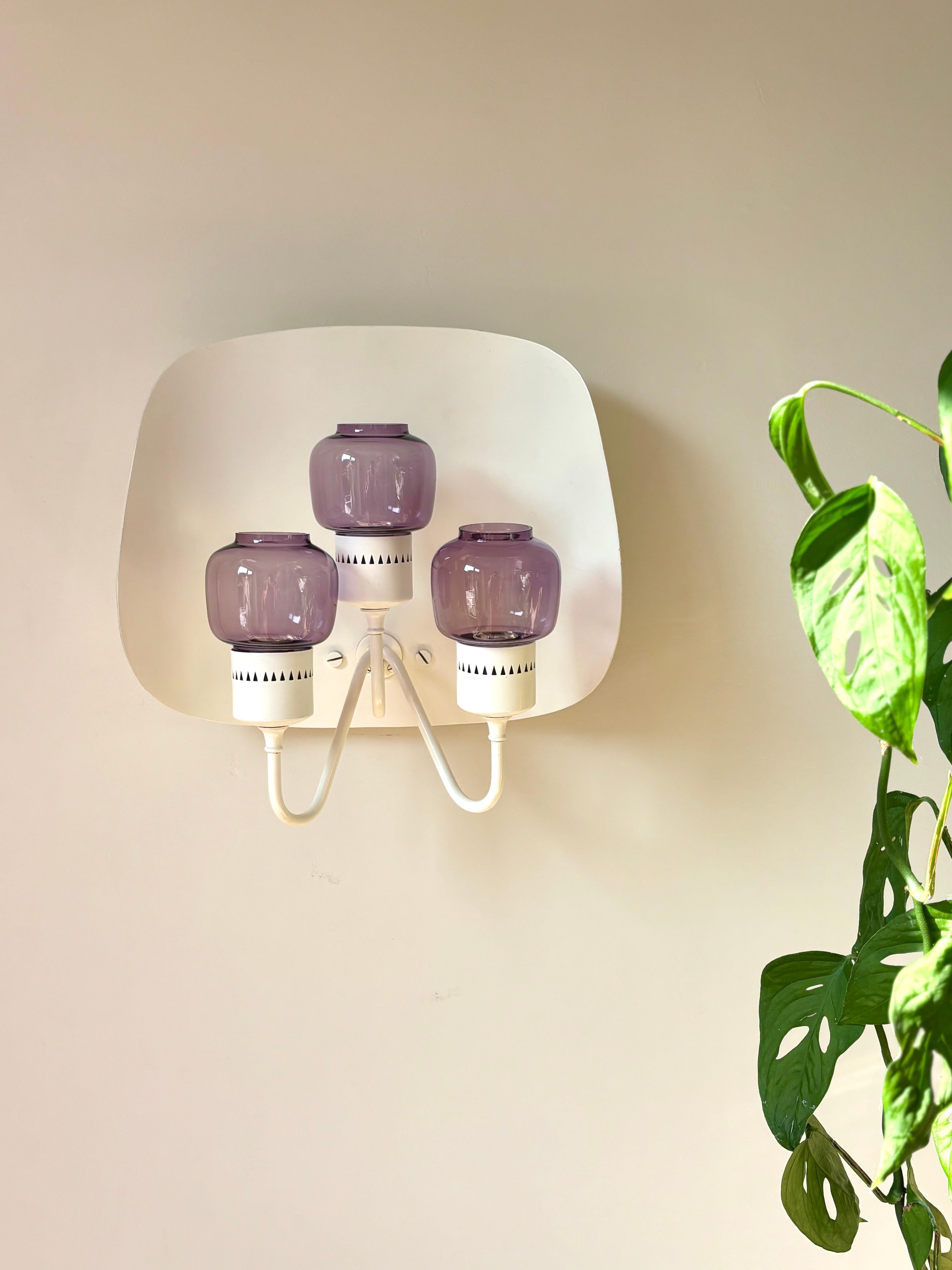 Applique murale The Modern Scandinavian Aginavian avec de rares abat-jours violets sur une monture en métal courbé laqué blanc chaud. Trois bras supportant chacun une ampoule E14 recouverte par de délicats abat-jour en verre aux formes douces.