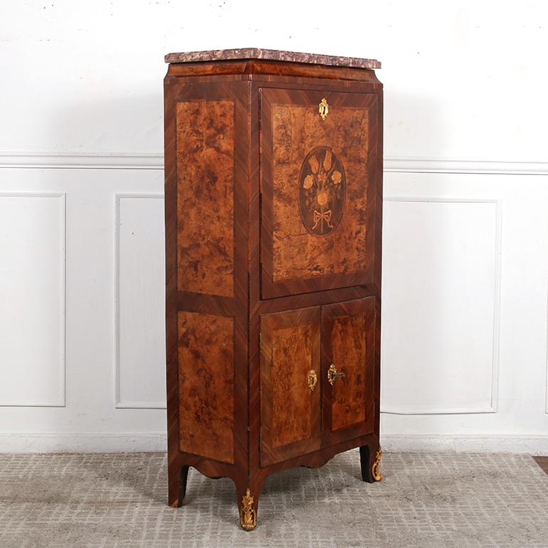Secrétaire Louis XV du XVIIIe siècle en palissandre et ronce de noyer, avec un plateau en marbre gris veiné. La façade est ornée d'une marqueterie de bois exotiques représentant un bouquet de fleurs noué par un ruban ; cette porte s'ouvre pour