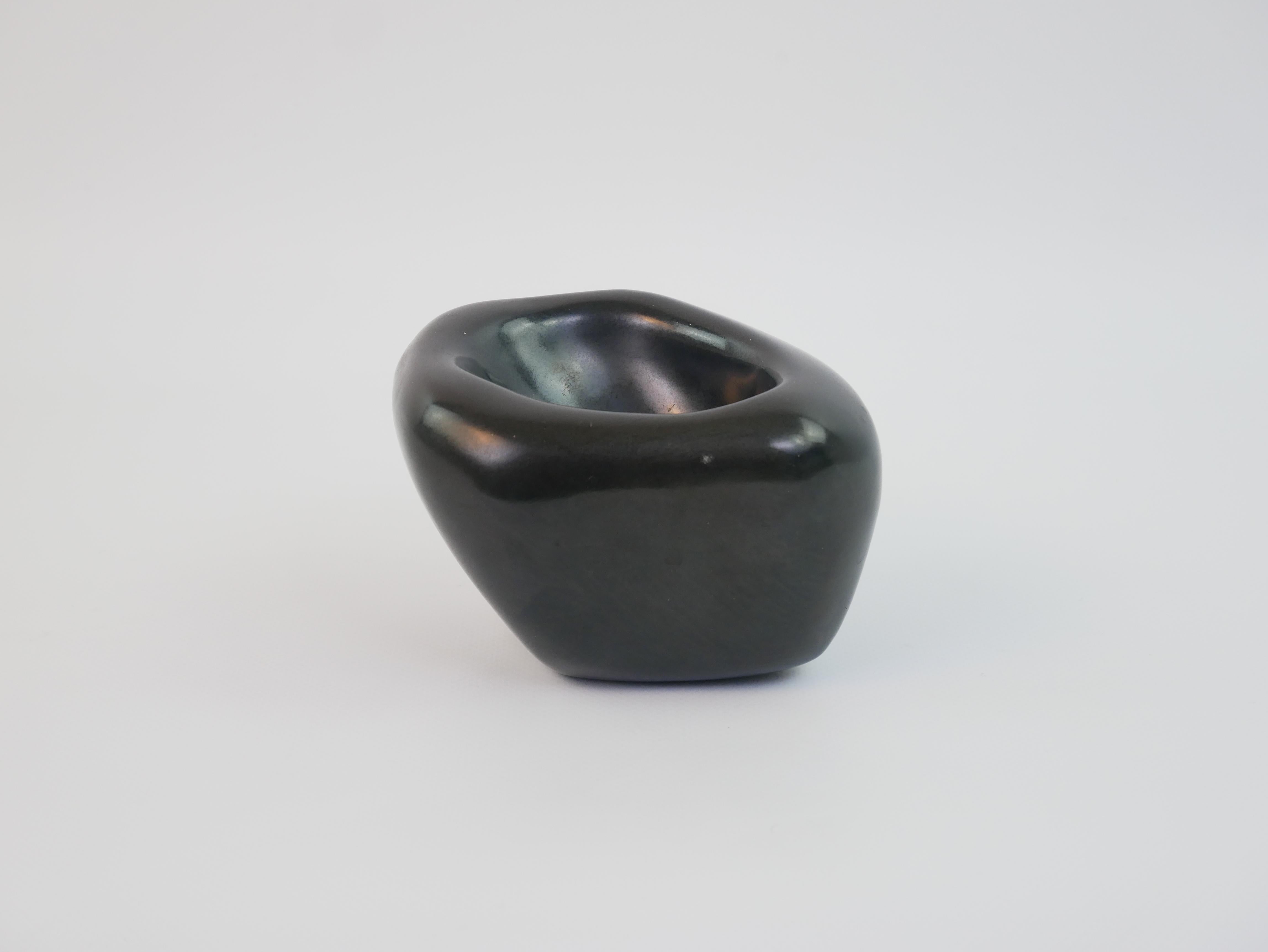 This small ceramic piece, likely a catchall or ashtray, embodies the elegant modernist style of Denise Gatard from the 1950s. Crafted in sleek black ceramic, it features an organic, asymmetrical form with a smooth, polished finish. The design is