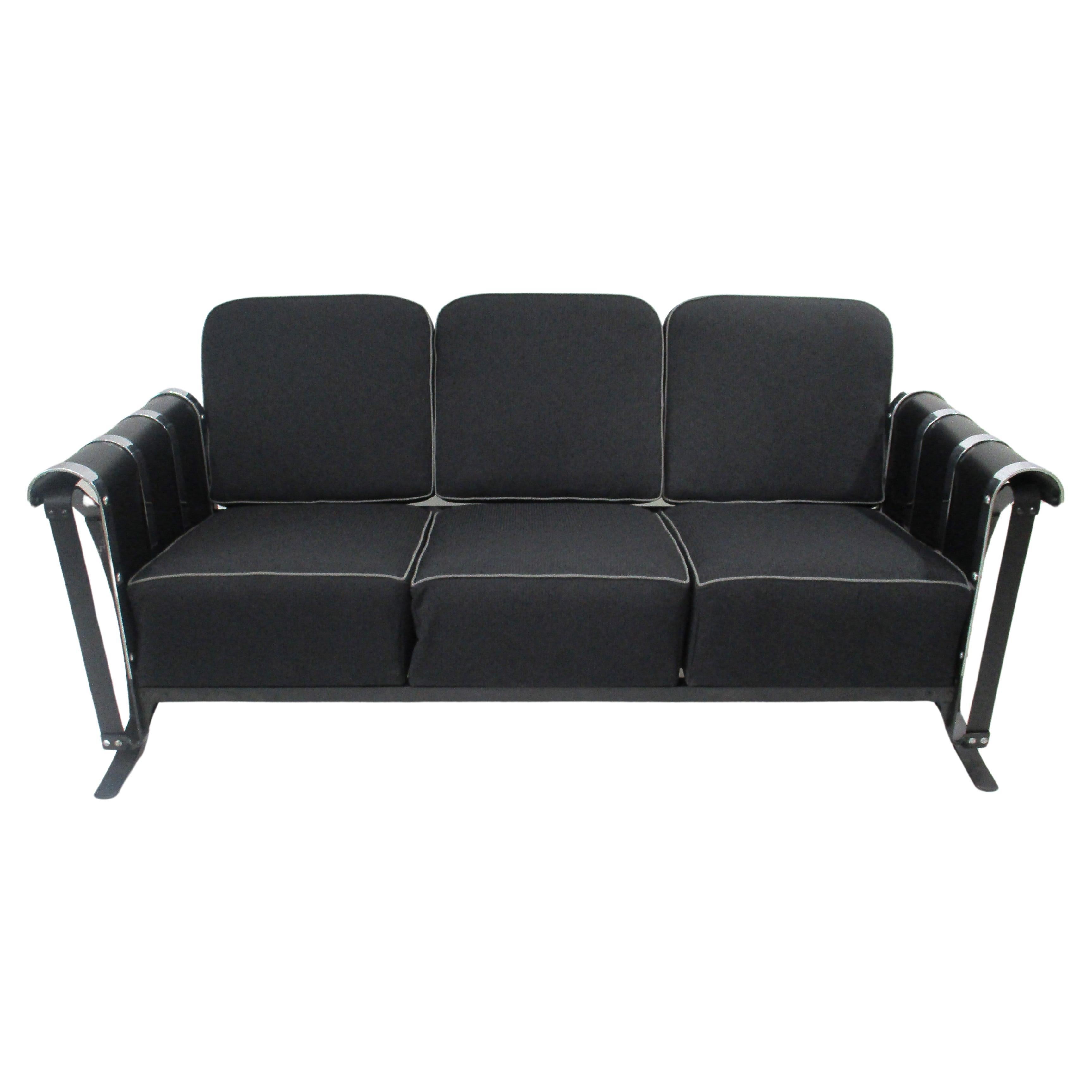 A steel framed satin black glider sofa with curved chrome and metal banded arms black upholstered cushions with medium gray piping . To each end there are sculptural flat bar supports integrated into the sides giving the piece an extra design