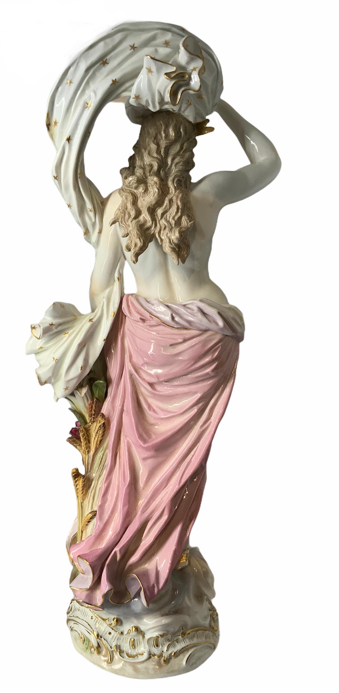 This Meissen porcelain represents a semi nude nymph with a gold crown standing up in victorious posture over a rocaille gilt round base. Beside her, there are a bunch of wheat spikes, iris and roses flowers. She features a bare torso and a pink and