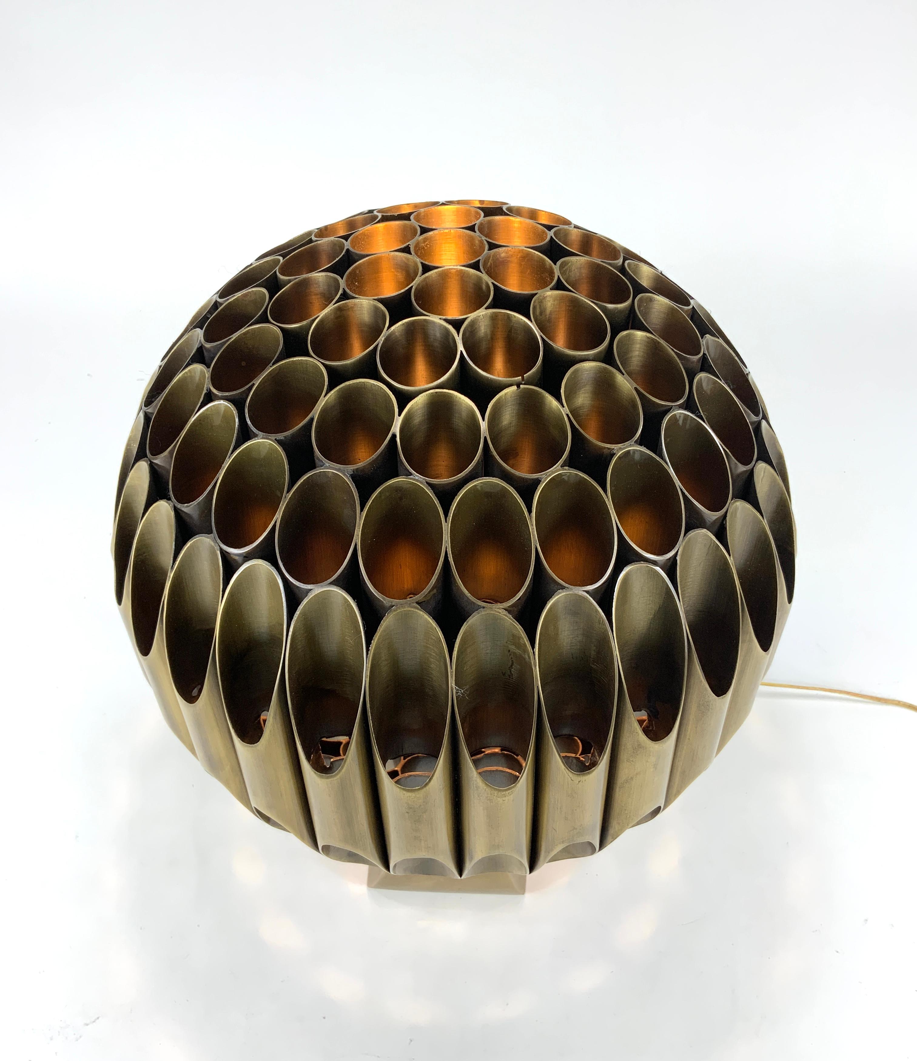 Michel Armand’s sculptural gilded bronze table lamp “Ruche” consists of metal cylindrical tubes that have been welded together and shaped into a perfect spherical form resting on an arched metal base.  A portion of the lamp is removeable from the