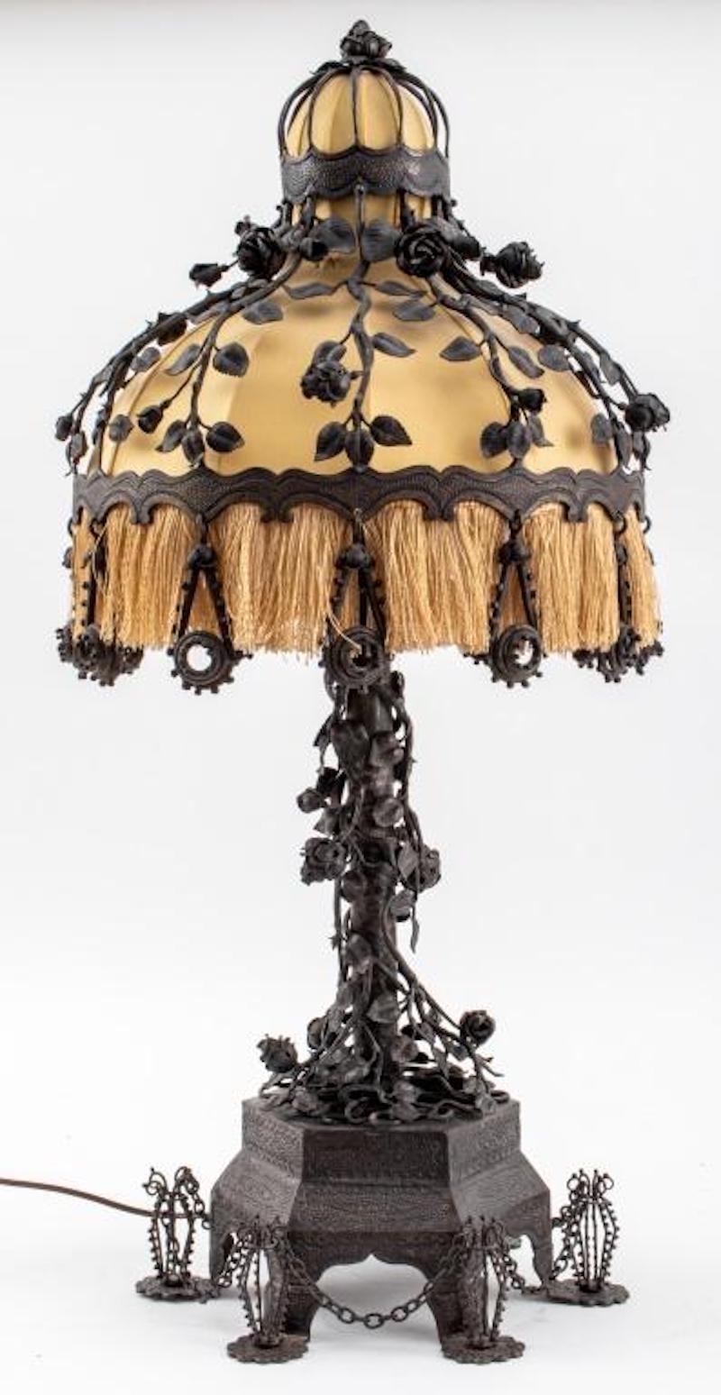 Beautiful craftsmanship bronze / wrought iron table lamp with rose flowers and thorns in high relief to the base and shade. The lamp features a bell shaped shade of beige fabric and fringe trim resting on an hexagonal base with geometric decoration