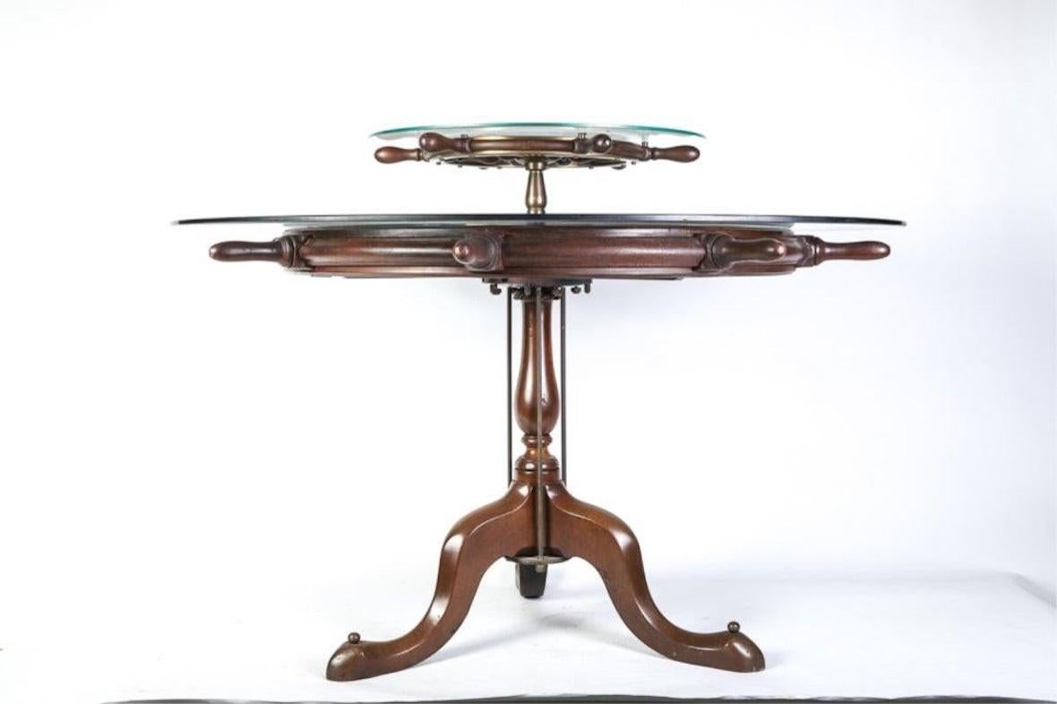 Ship's wheel stamped WWII US Navy with brass mounts adaptively reused as a tabletop for a Queen Anne base with a vasiform post and (3) cabriole legs. A second ships wheel serves as a Lazy Susan. (2) glass tops nest into wheel rabbit. The feet are