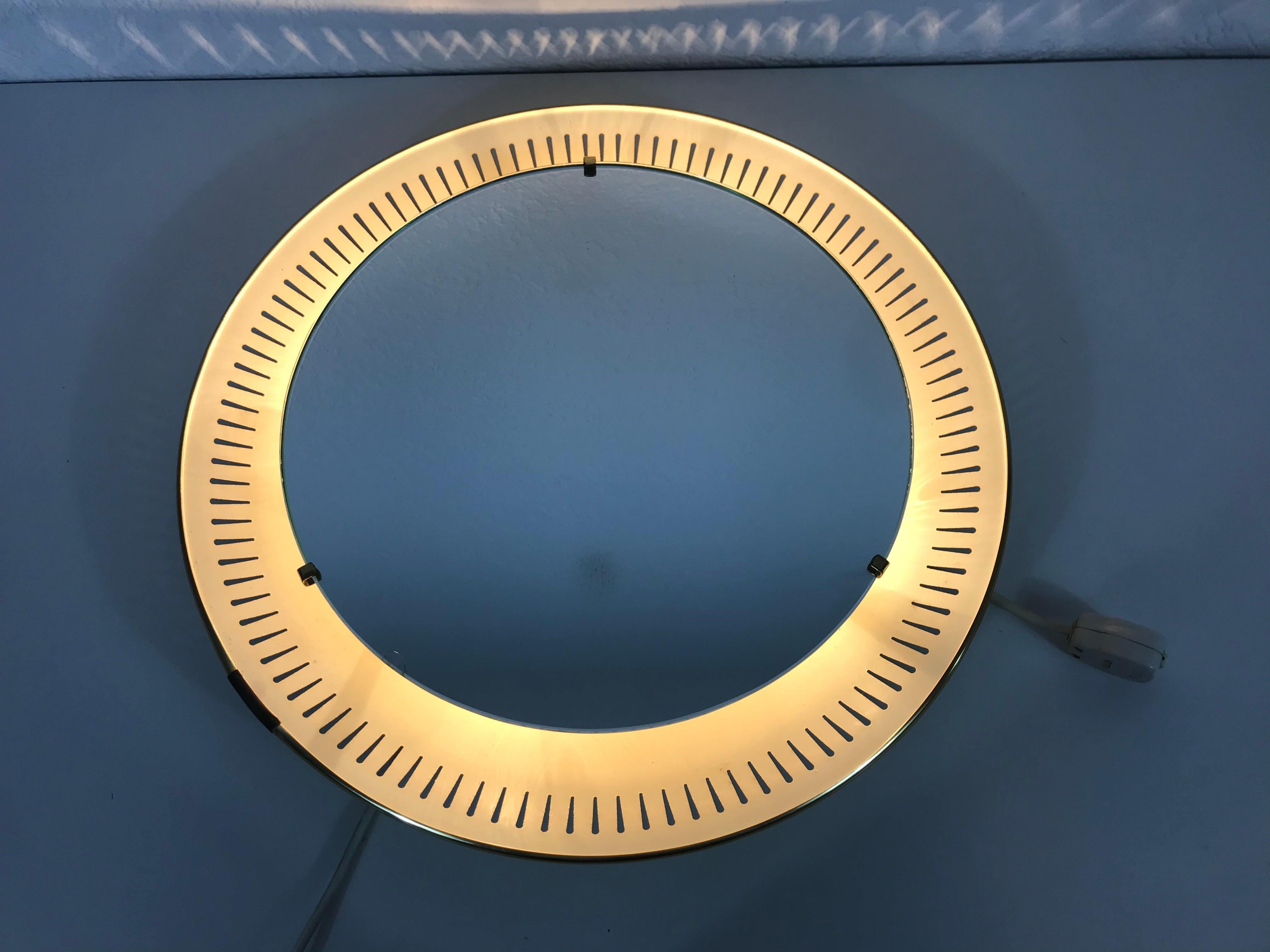 A Hillebrand illuminated wall mirror from the 1950s made in Germany. The mirror has a round metal design. There are E14 light bulbs inside the frame. 3 brass screws which secure the glass to the aluminium frame. The mirror is in a good vintage