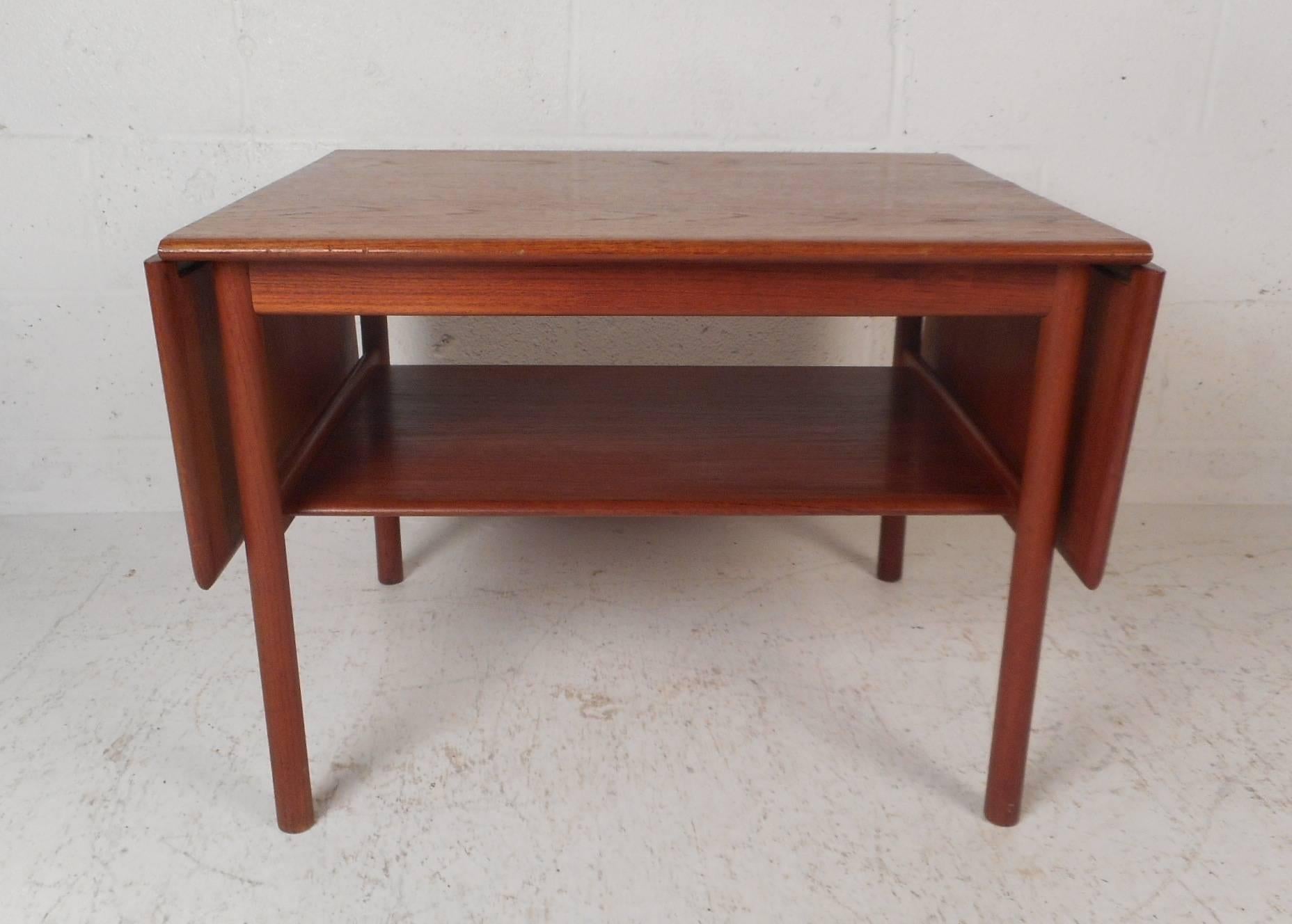 This stunning vintage modern coffee table features a drop-leaf design extending the width all the way to 48 inches. The perfect space saving table with a lower shelf for additional storage and an elegant teak wood grain throughout. This unique