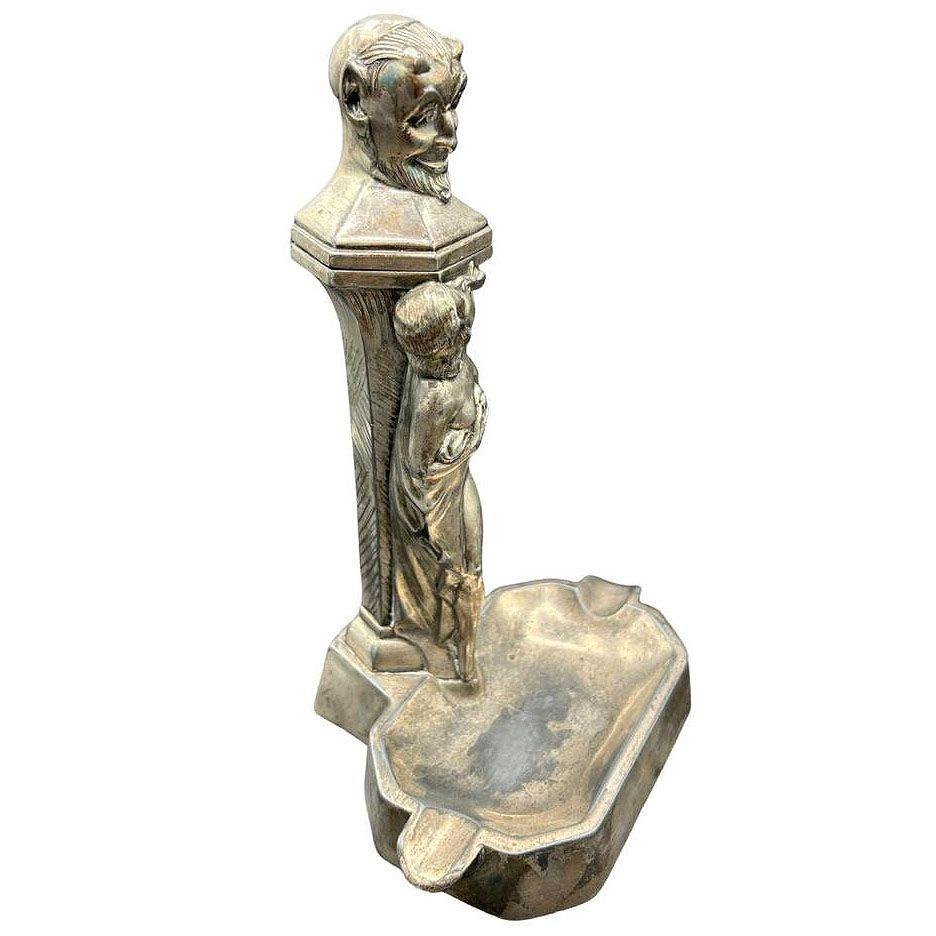 Questo accendino e posacenere da tavolo in metallo bianco degli anni '20 presenta un busto di Diavolo/Satyr sopra una ninfa fumatrice con i seni nudi scoperti che riprende la storia della tentazione nel giardino dell'Eden, sostituendo una mela con