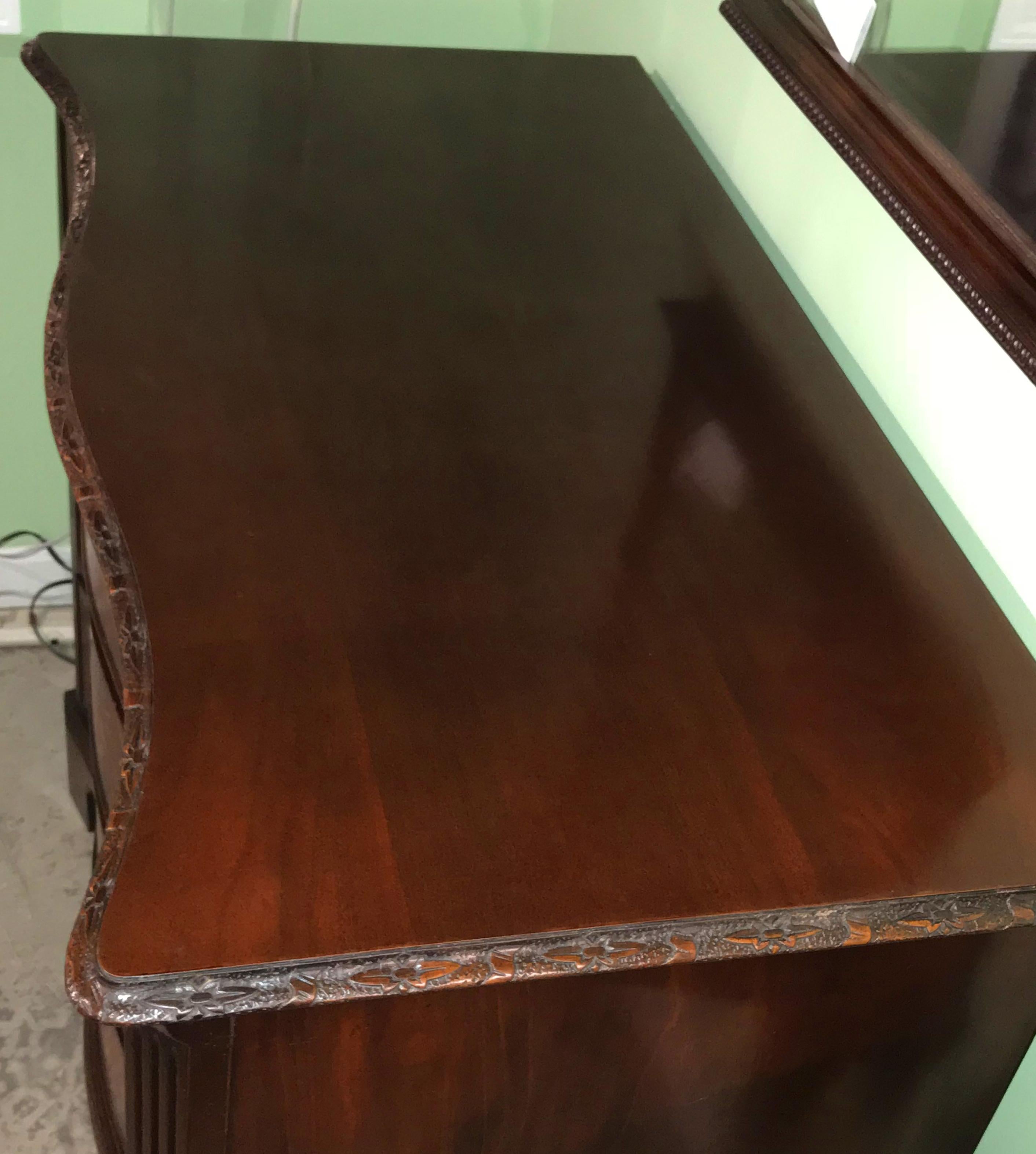 A beautiful mahogany serpentine chest of drawers in the Chippendale style, made by Old Colony Furniture Company, with conforming top featuring detailed foliate and ribbon carving on its edge, surmounting four drawers with bookmatched crotched