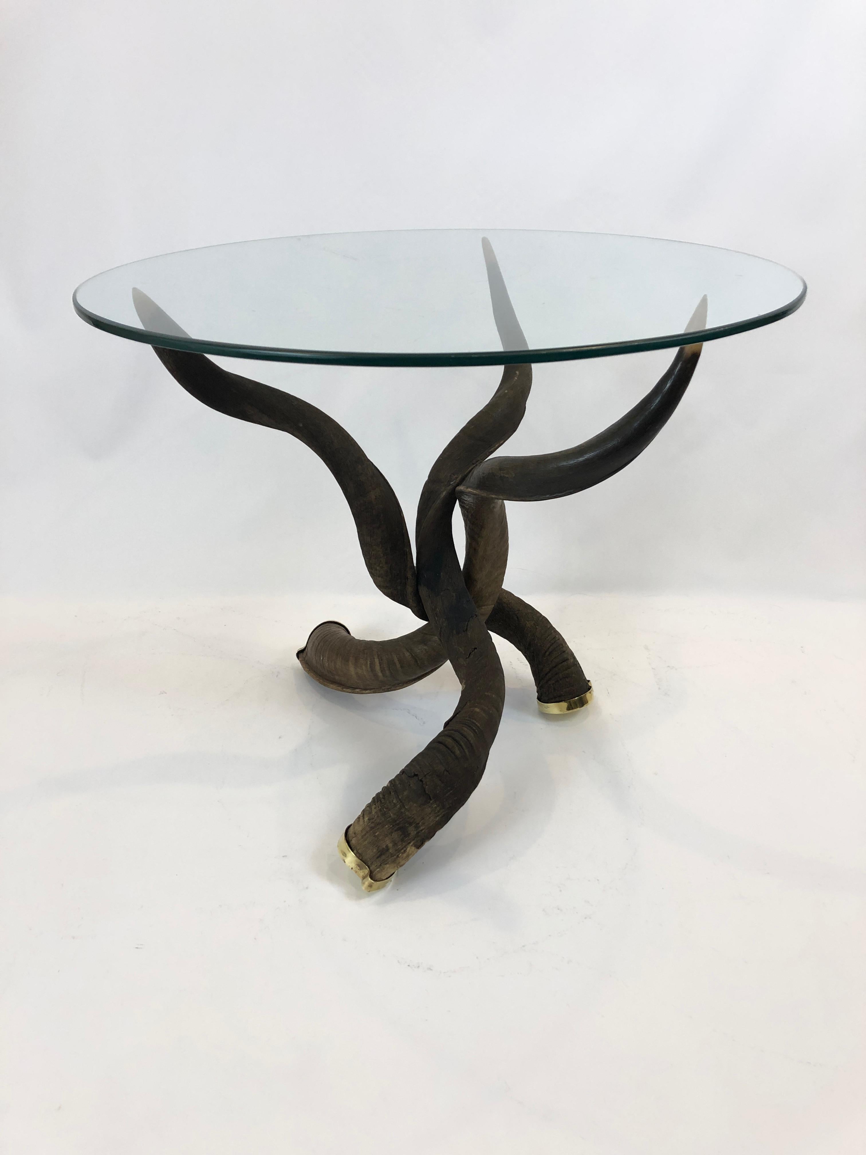 A sculptural custom made tripod coffee table having 3 kudo horns elegantly arranged with tips at the top perfectly poised to hold the brand new bevelled round piece of glass. The ends of the horns on the floor terminate in brass caps. Base is 21 W