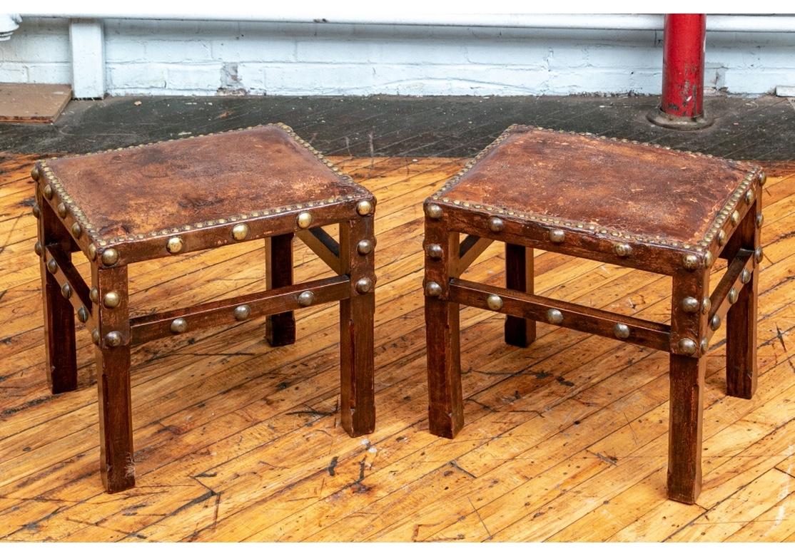 The small rectangular benches with old brown leather seats and brass nail heads around the top edges. The wood frames with carved legs with canted edges and square stretchers on all sides. The frames mounted overall with brass bosses (lacking one on