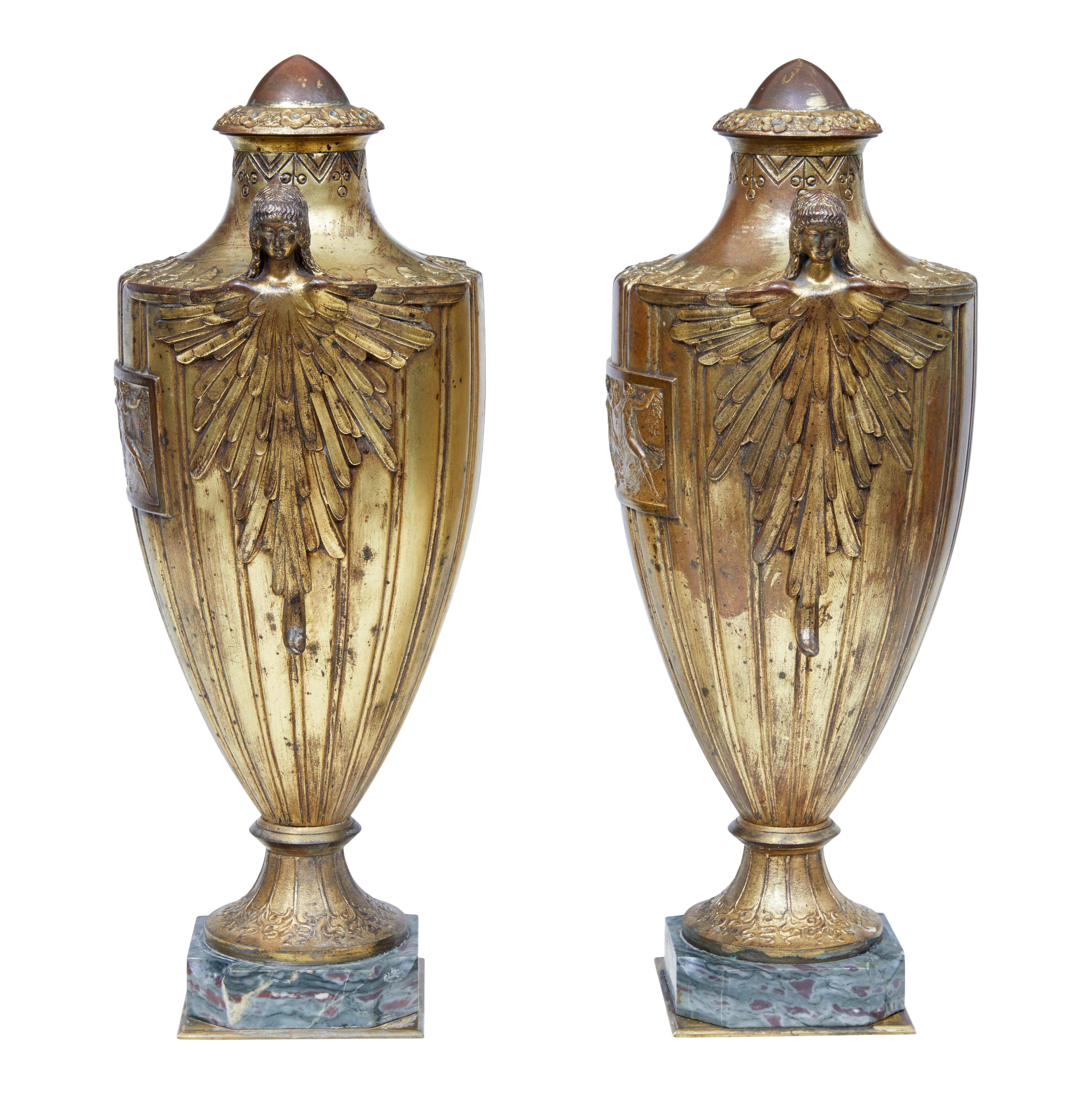 Opportunity to own a fine pair of period art deco urns, circa 1920.

Solid bronze with channeled edge which flows down to the base. Beaked top finial surrounded by cast flowers with central glass detailing. Winged female figures on both sides. On