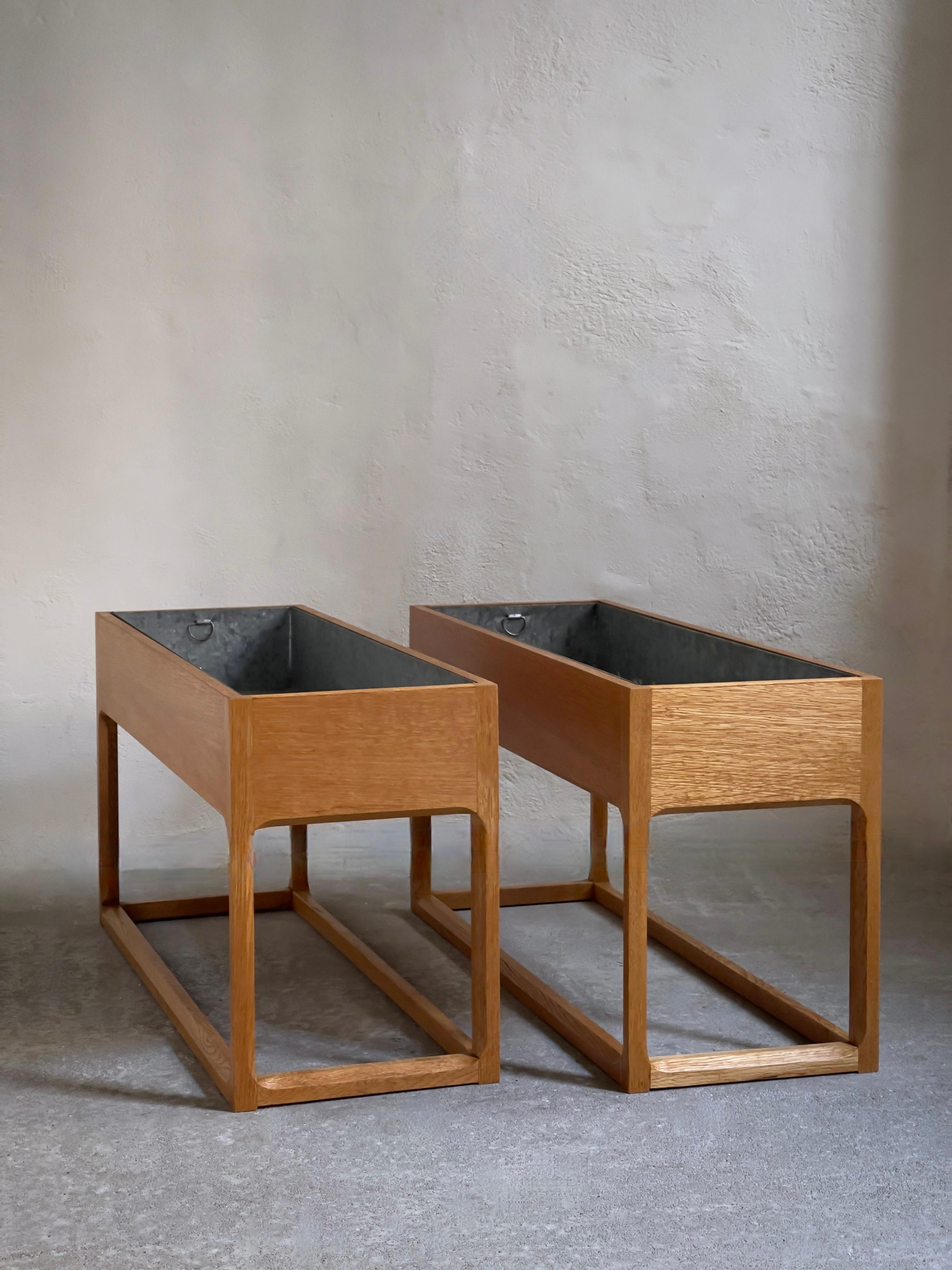 Presenting a captivating piece of Danish craftsmanship from the 1960s—a rare pair of indoor planters or jardinieres skillfully crafted by the renowned cabinetmaker Axel Kjersgaard. 

Crafted from oak, a traditional and durable hardwood, these