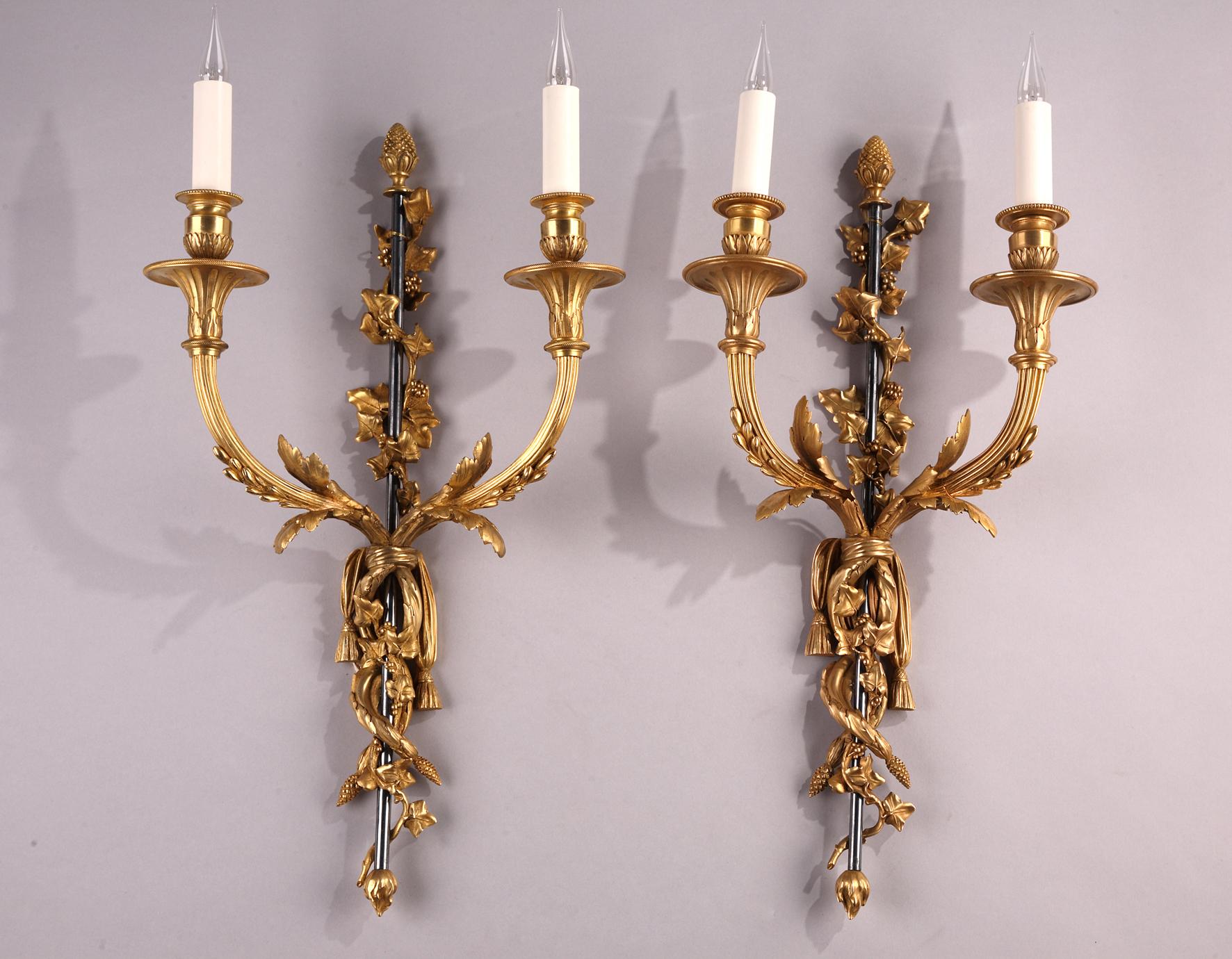 Chased and gilt bronze sconces attributed to H. Vian, each comprising a “gun barrel” shaft surmounted by a pine cone and entwined with ivy branches. Two foliated creepers tied with a cloth are interlaced around the central shaft and give rise to two