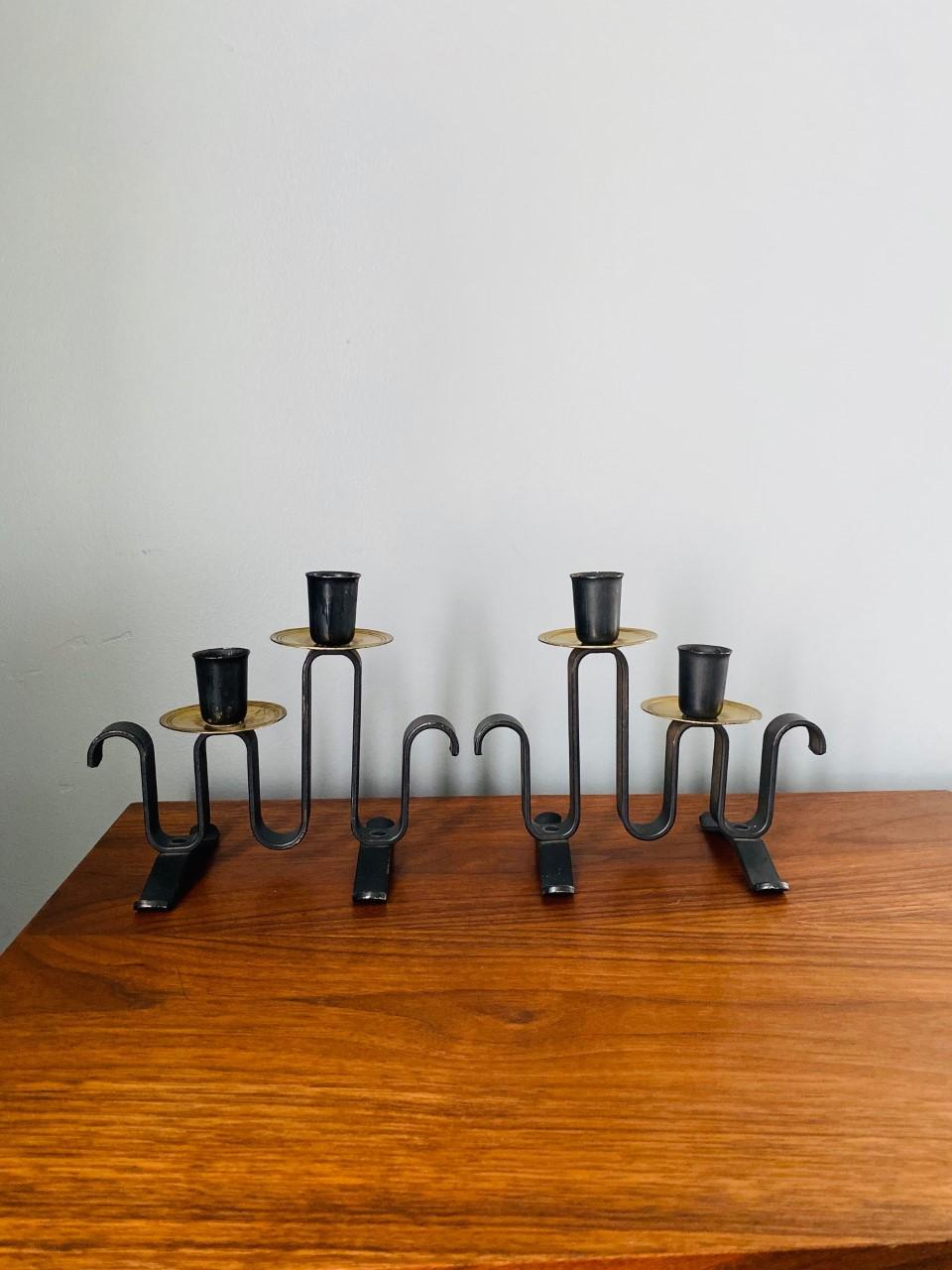 Rara coppia di candelabri postmoderni in ferro battuto. I due pezzi sono grafici e dalle linee pulite. Il ferro battuto a forma di onda ha come apice un disco dorato che sostiene il cero della candela, mentre le due gambe garantiscono una stabilità