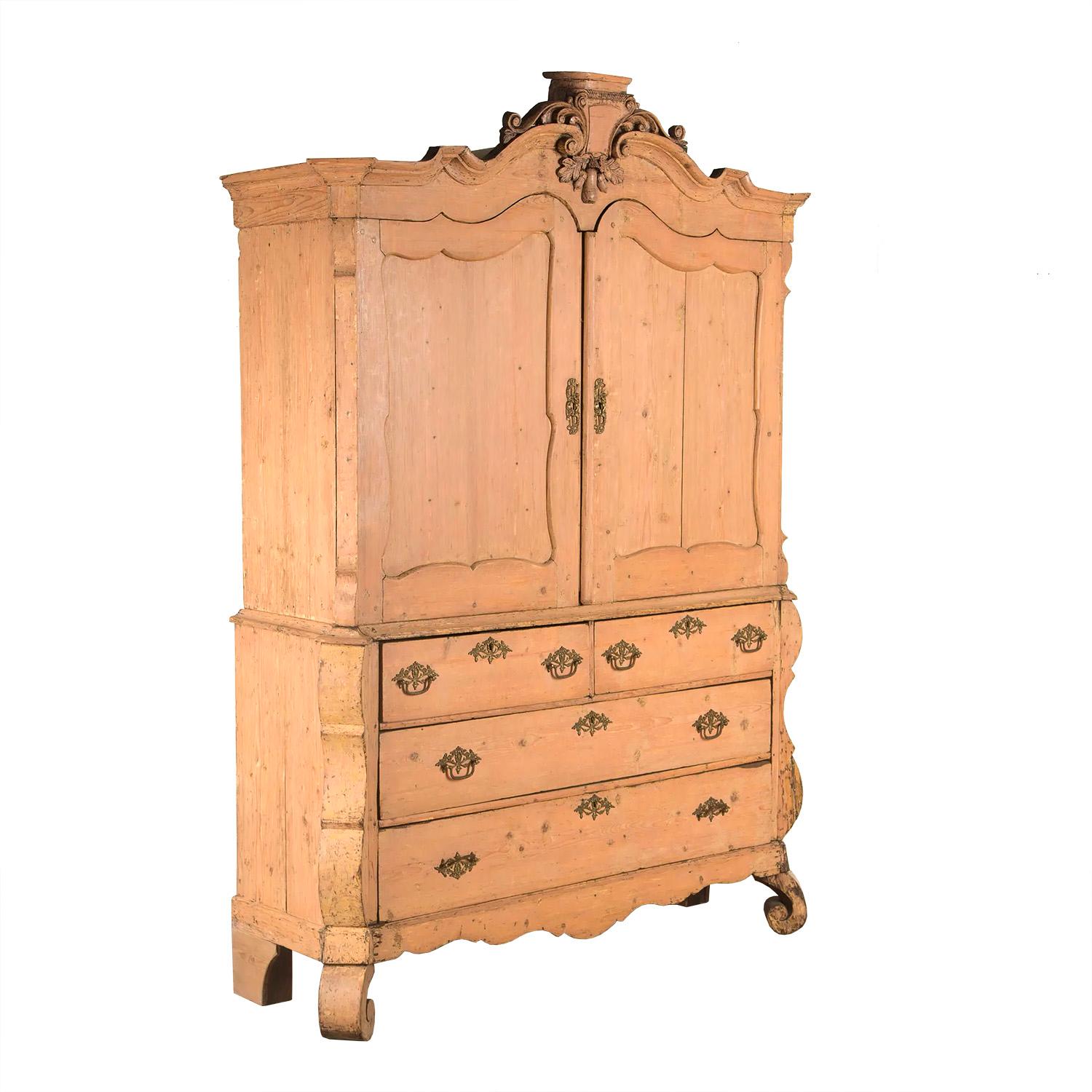 A two part Dutch country pine cabinet with decorative carved pediment featuring a cartouche and acorns with leaves. This piece features two doors opening to storage shelves with small drawers. The bottom section has two long and one two part