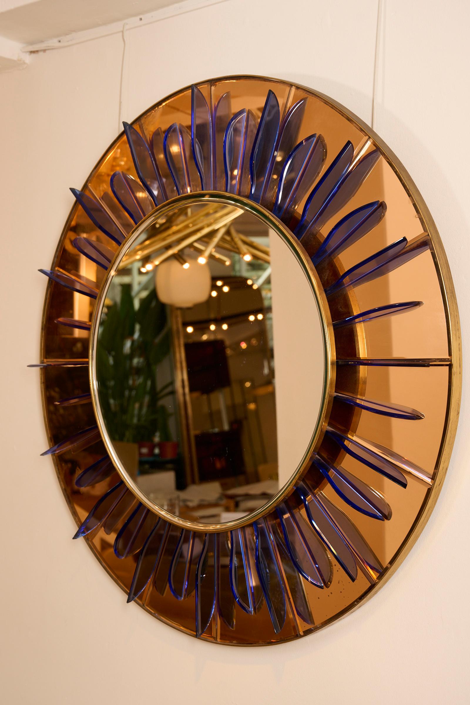 Impressive circular mirror with bevel in brass frame mounted on a peach/bronze glass with cut blue glass blades radiating from the mirror.

