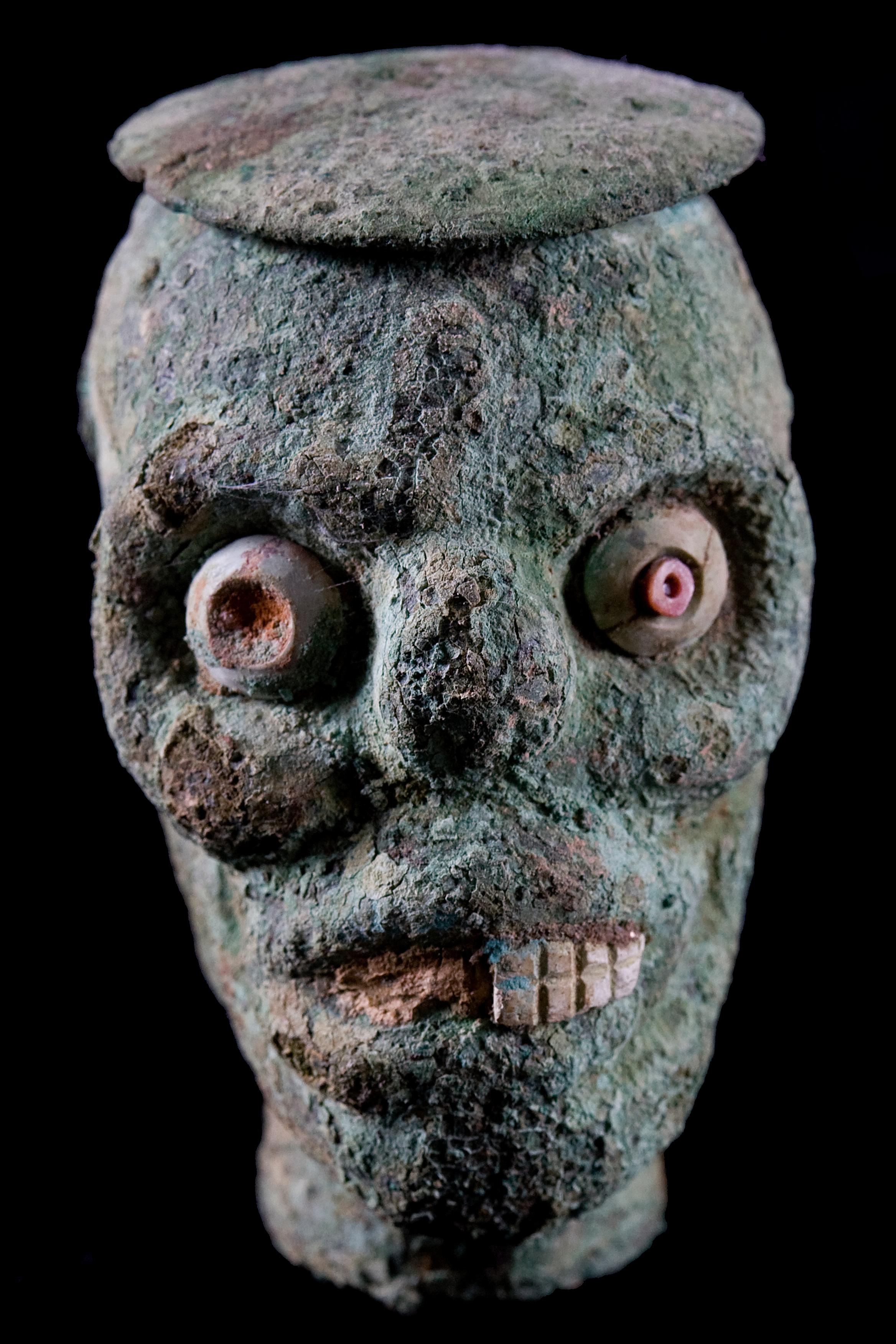 A hollow molded copper skull head, lidded vessel having well defined relief features with inset shell eyes and teeth. Lid has a double headed applicator. Overall deep blue-green patina. Double perforations on both sides of the rim. Intact other than
